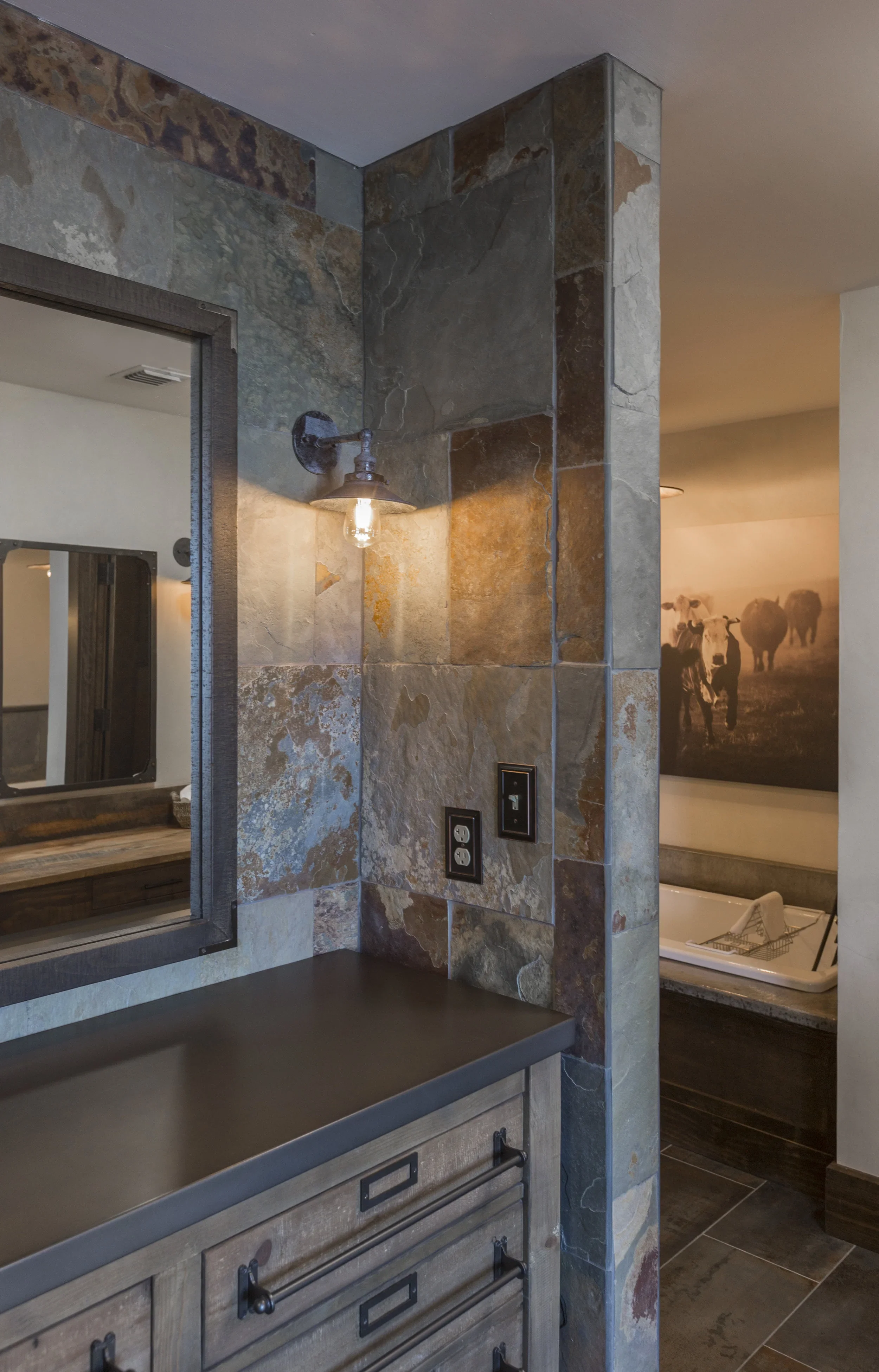 A rustic bathroom vanity with a black countertop, a large mirror, a vintage-style light fixture, stone tile walls, and a photo of cows with a bathtub in the background.