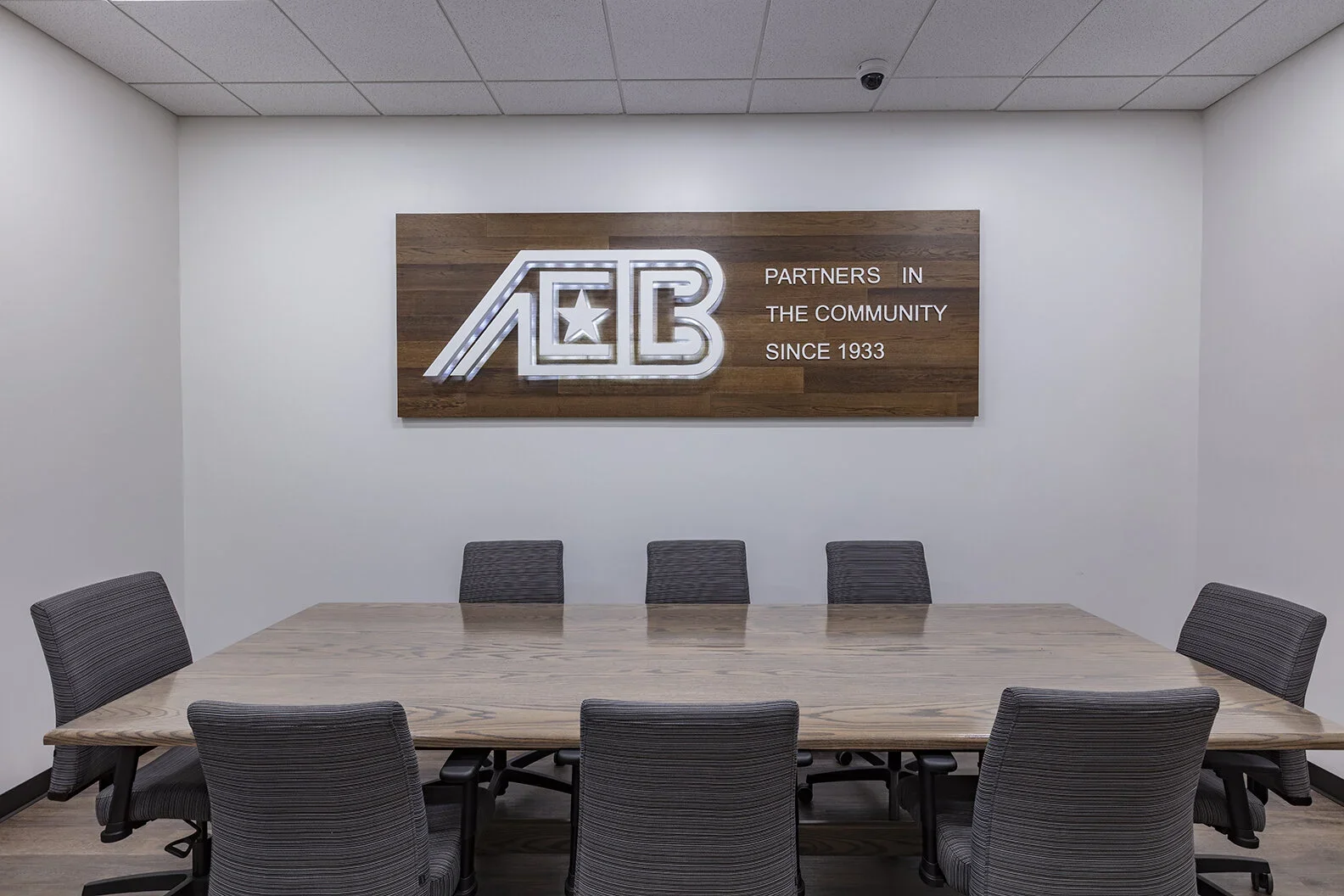 A conference room with a wooden table and eight gray upholstered chairs, six on one side and one on each of the other sides. On the wall behind the table, there is a wooden panel with a backlit logo and the text "Partners in the Community Since 1933"