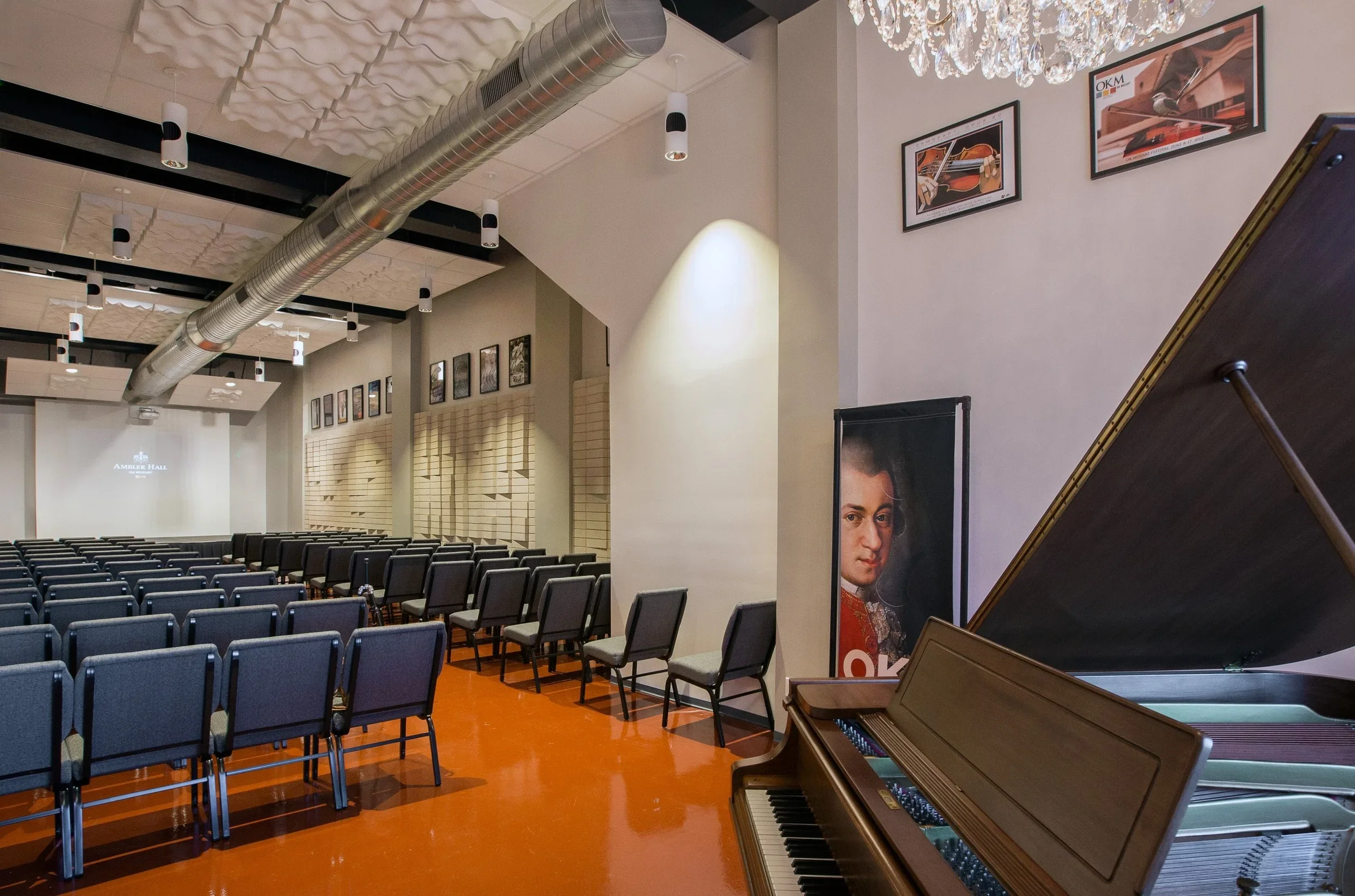 An auditorium with rows of gray chairs, a grand piano near the right side, framed pictures on the wall, a chandelier, and a projector screen at the front displaying 'Ambler Hall' in a modern venue.