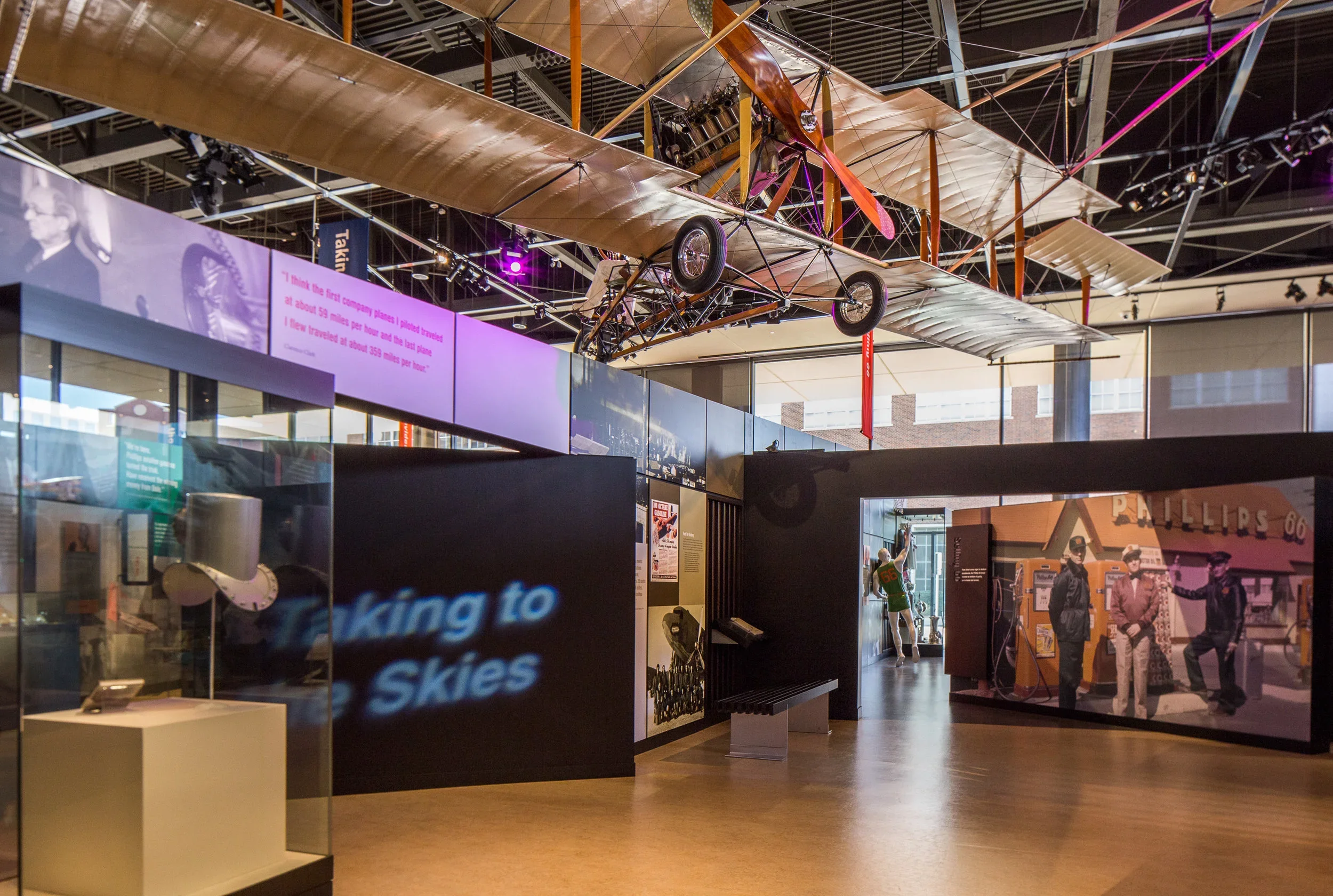 Interior of a museum exhibit featuring an antique airplane suspended from the ceiling, with display cases and images related to aviation history on the walls and floor.
