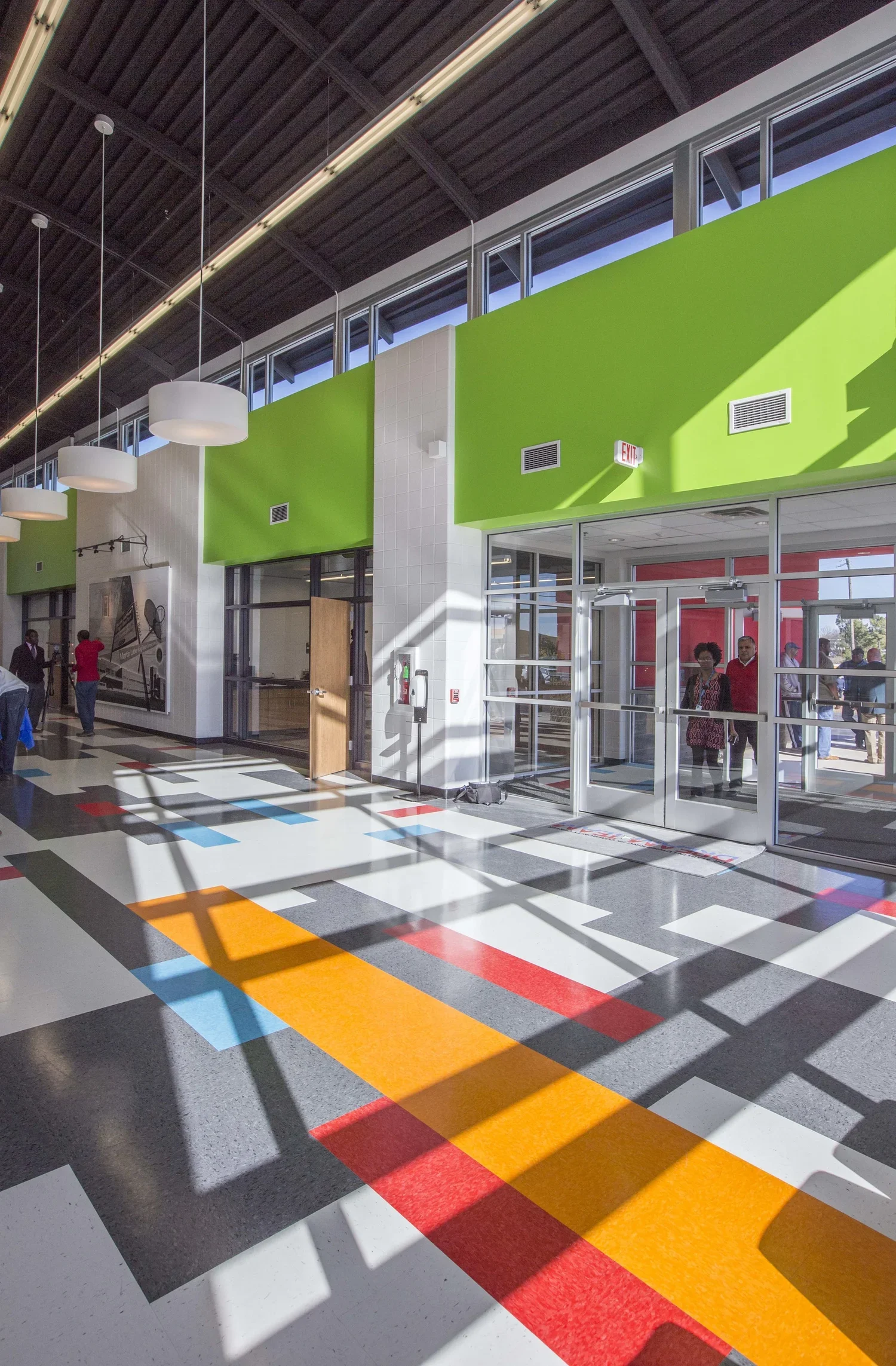 Interior of a modern building with colorful striped flooring, white and green walls, large windows, and hanging ceiling lights. Several people are walking or standing near the entrance and inside.