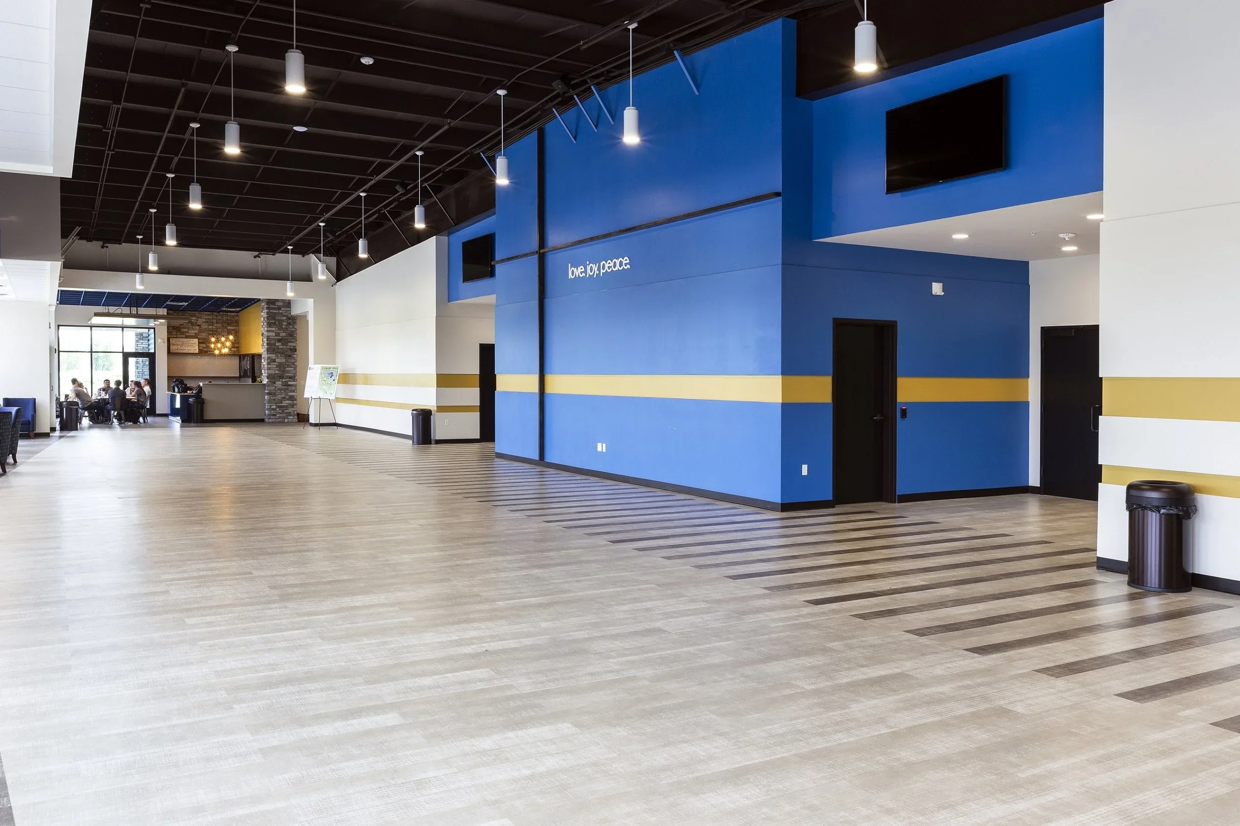 Empty lobby area inside a modern building with a blue wall, black doors, striped yellow and white wall accents, a digital screen, and a small seating area with tables and chairs near large windows.