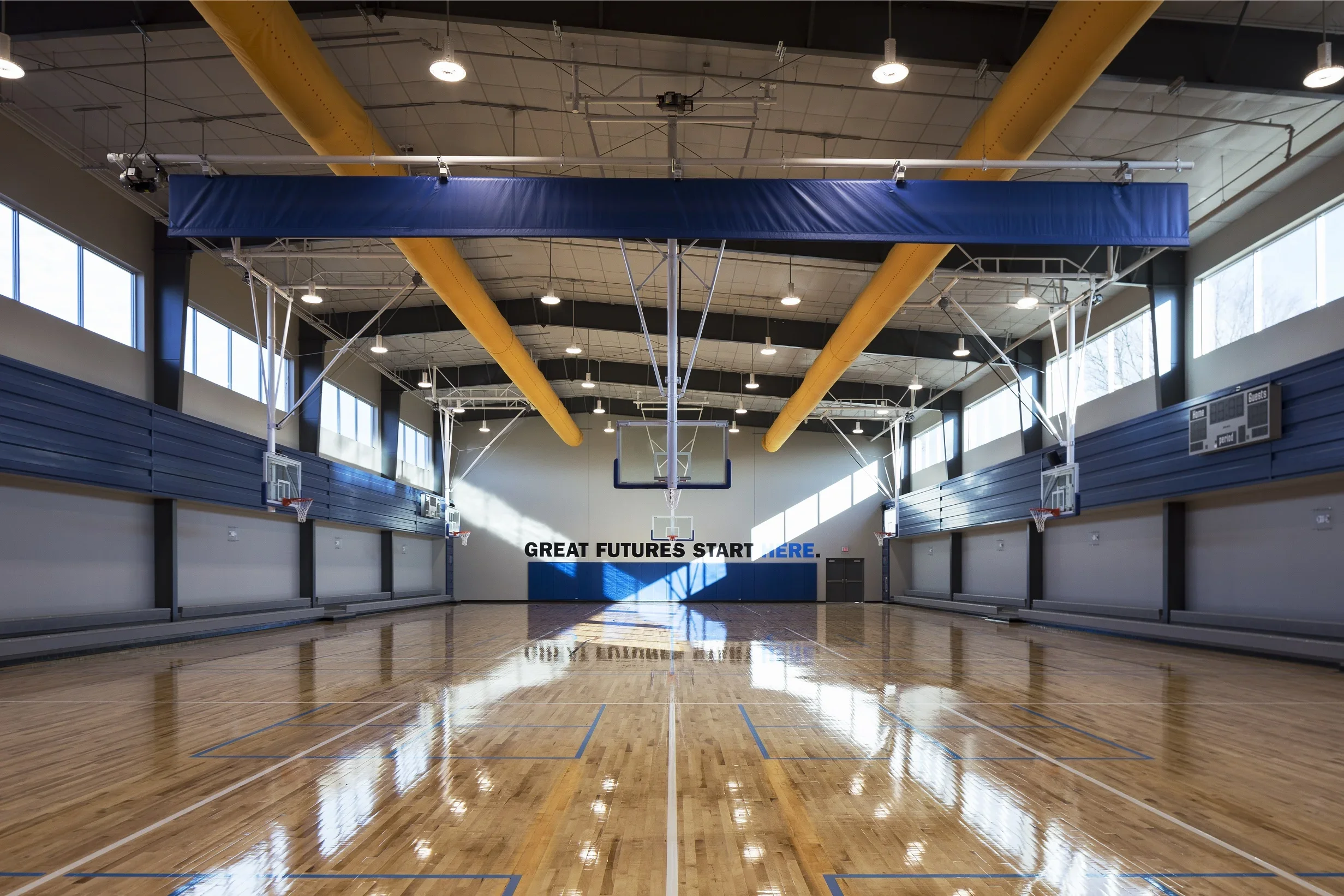 Empty indoor basketball court with polished wooden floor, multiple basketball hoops, and large windows letting in natural light. A motivational sign on the far wall reads 'Great futures start here.' Suspended yellow ducts and ceiling lights are visib