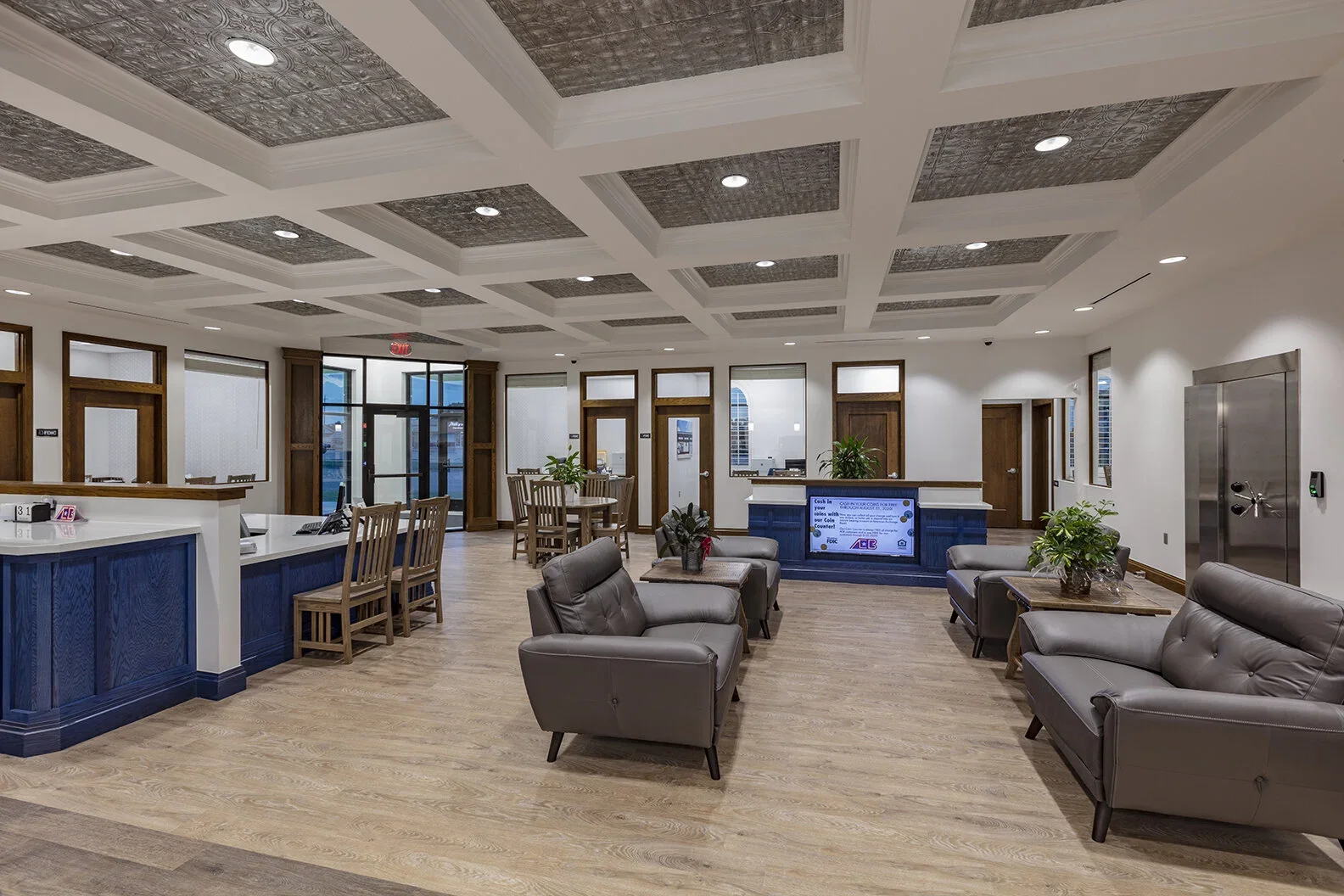 A lobby with a reception desk, seating area with leather chairs and sofas, wooden tables with plants, and a view of some doors and windows, illuminated by recessed ceiling lights.