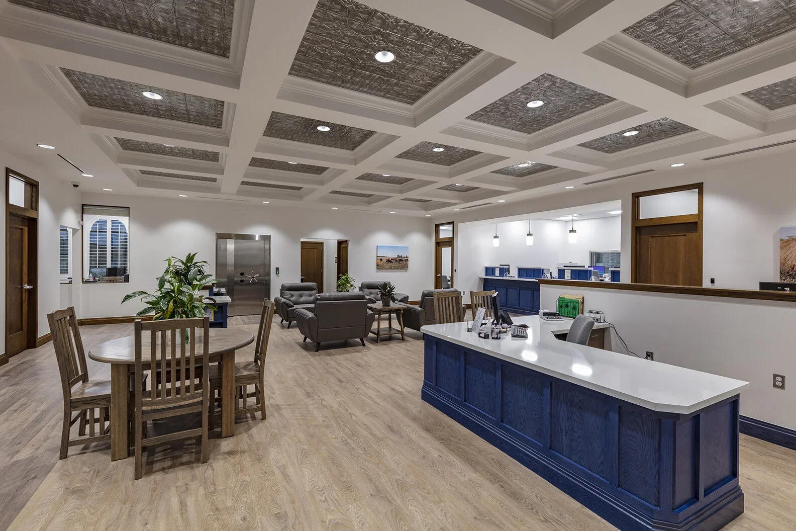 A spacious lobby with a high, coffered ceiling and recessed lighting. The space includes a round wooden table with four chairs to the left, a seating area with gray sofas and armchairs in the center, and a white reception counter with a blue base on 