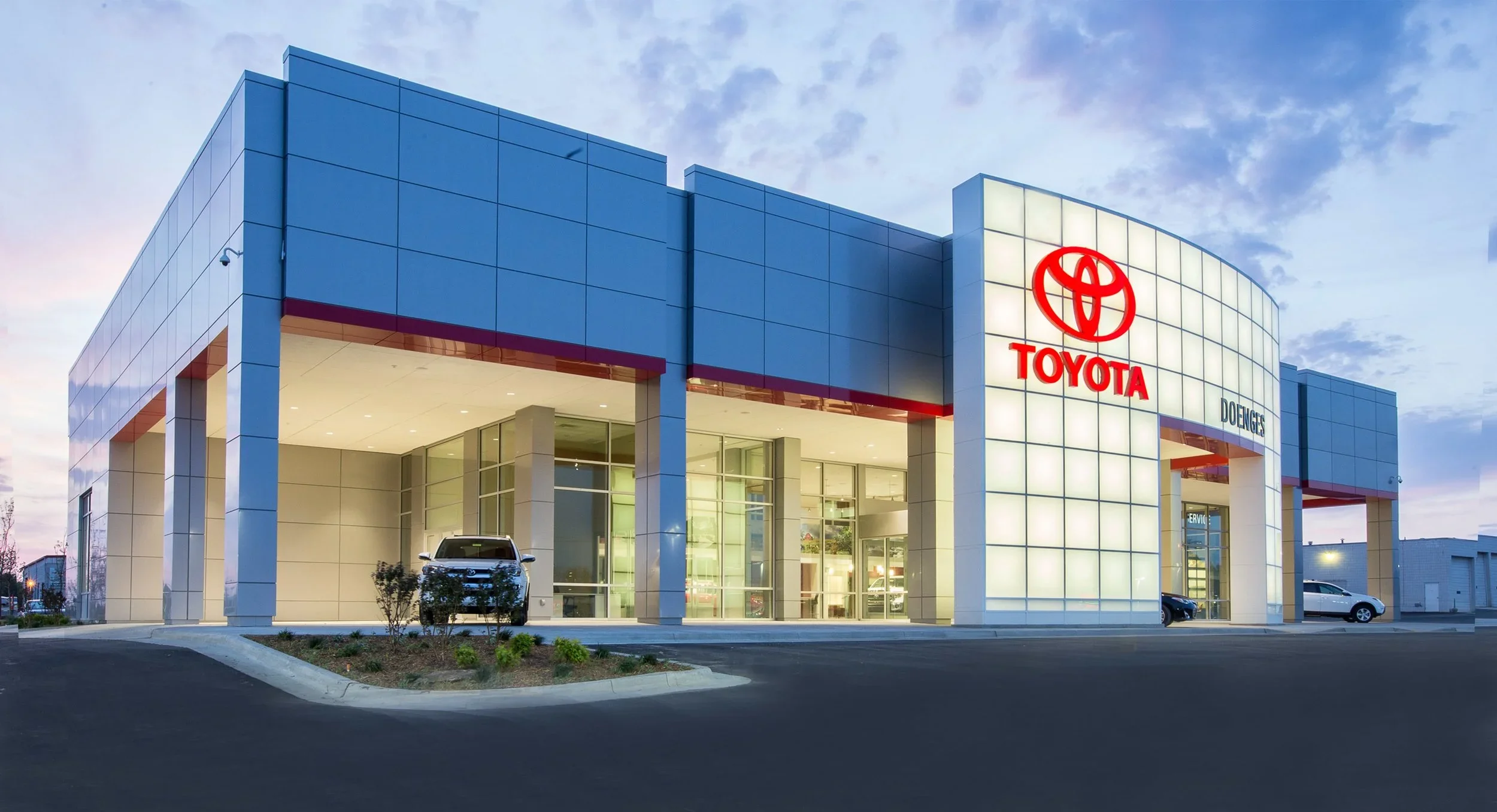 Modern Toyota car dealership building with illuminated logo at dusk, featuring blue panels and a white front with large glass windows and cars parked outside.