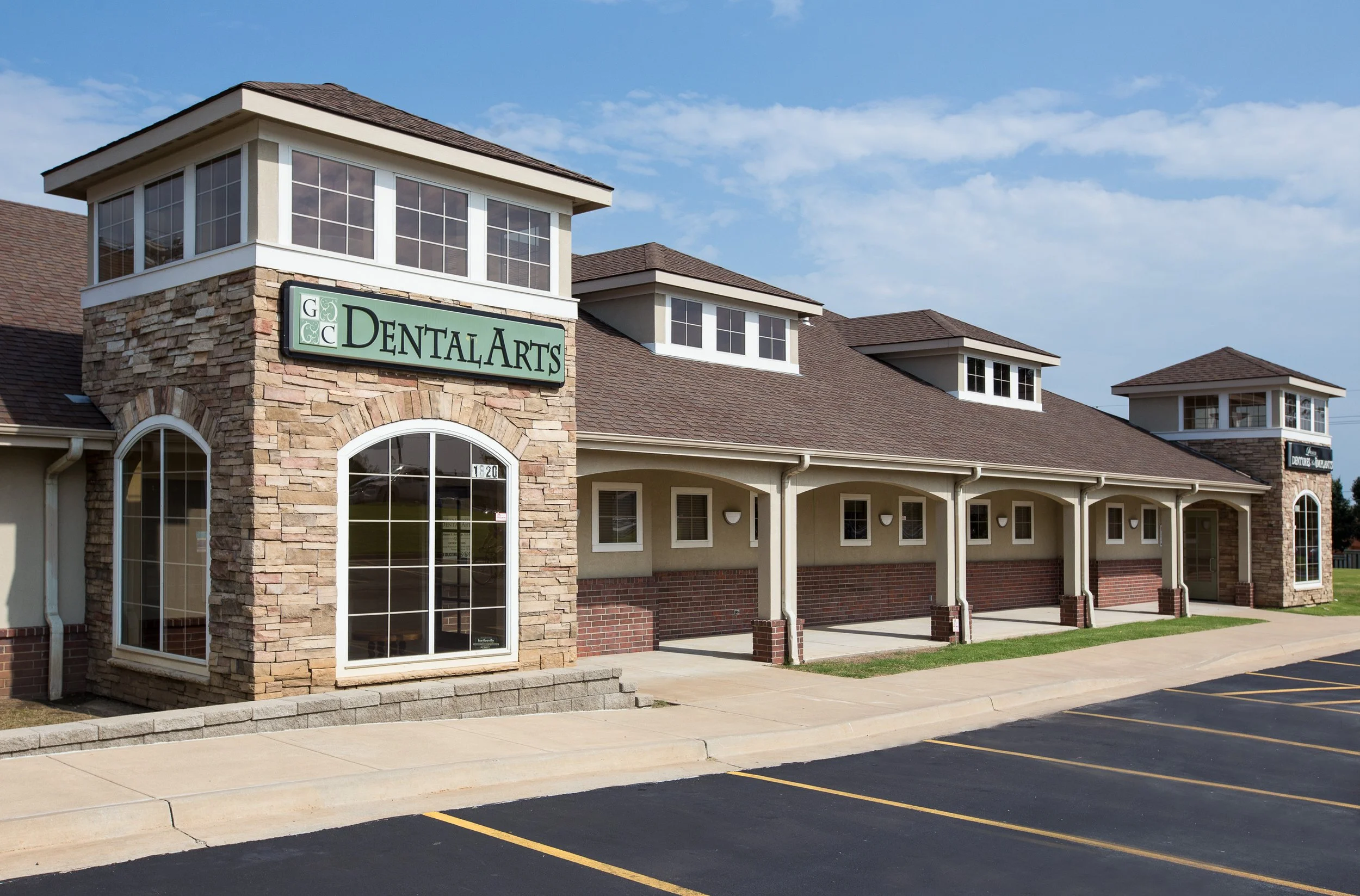Exterior view of a dental office building with a sign that reads 'Dental Arts', featuring a stone facade, multiple windows, and wooden posts in front of a parking lot.