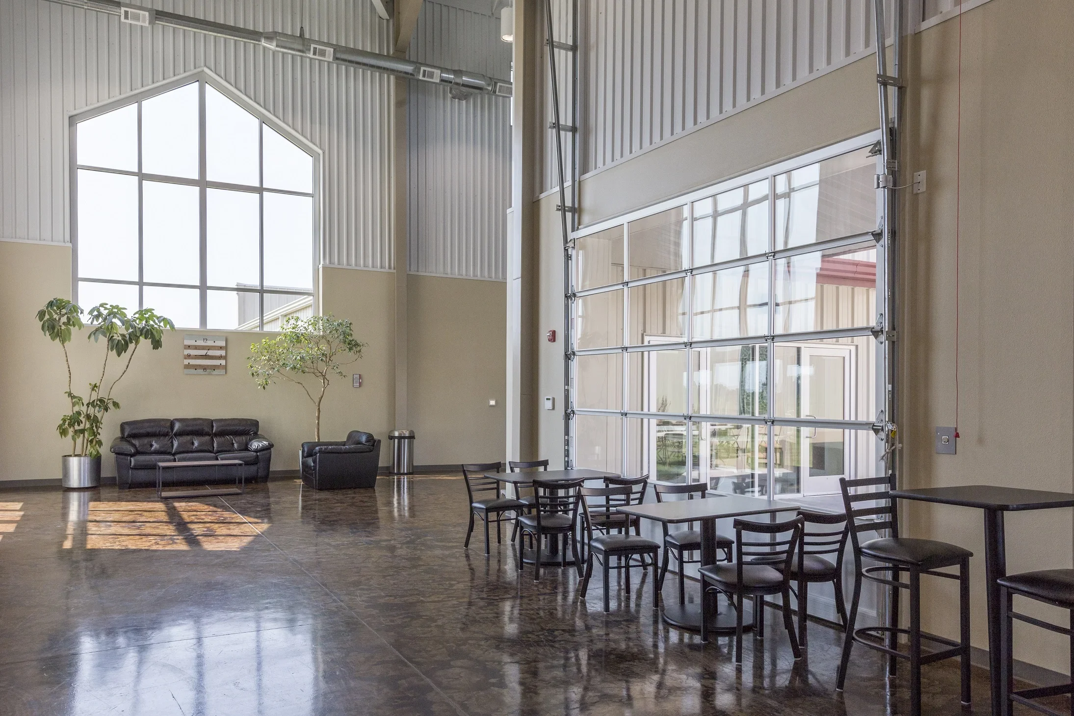 Interior of a spacious lounge area with a large glass garage door, black leather sofa, side armchair, two indoor trees, and tables with chairs, featuring polished concrete floors and high ceilings.