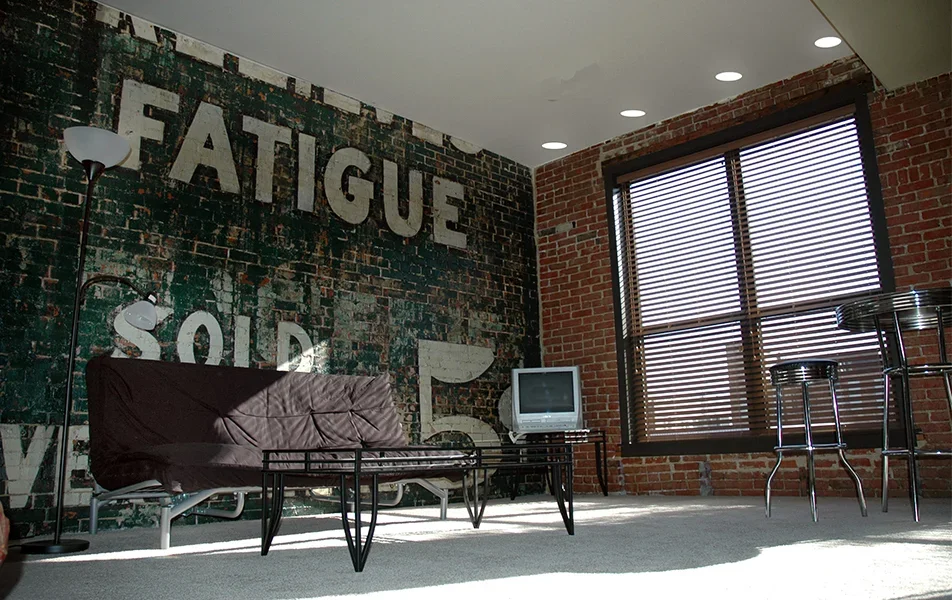 Interior of a loft-style living room with a brick wall, a large window with blinds, a brown sofa with a cover, a small TV on a stand, a tall floor lamp, and bar stools around a high table, with a sign on the brick wall reading 'FATIGUE SOLD'.