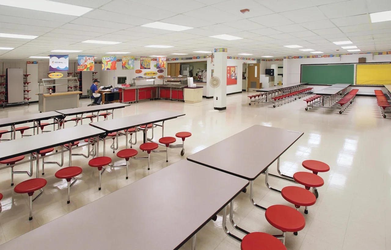 Empty school cafeteria with rows of red-stool-topped tables, a chalkboard at the back, and colorful banners hanging from the ceiling.