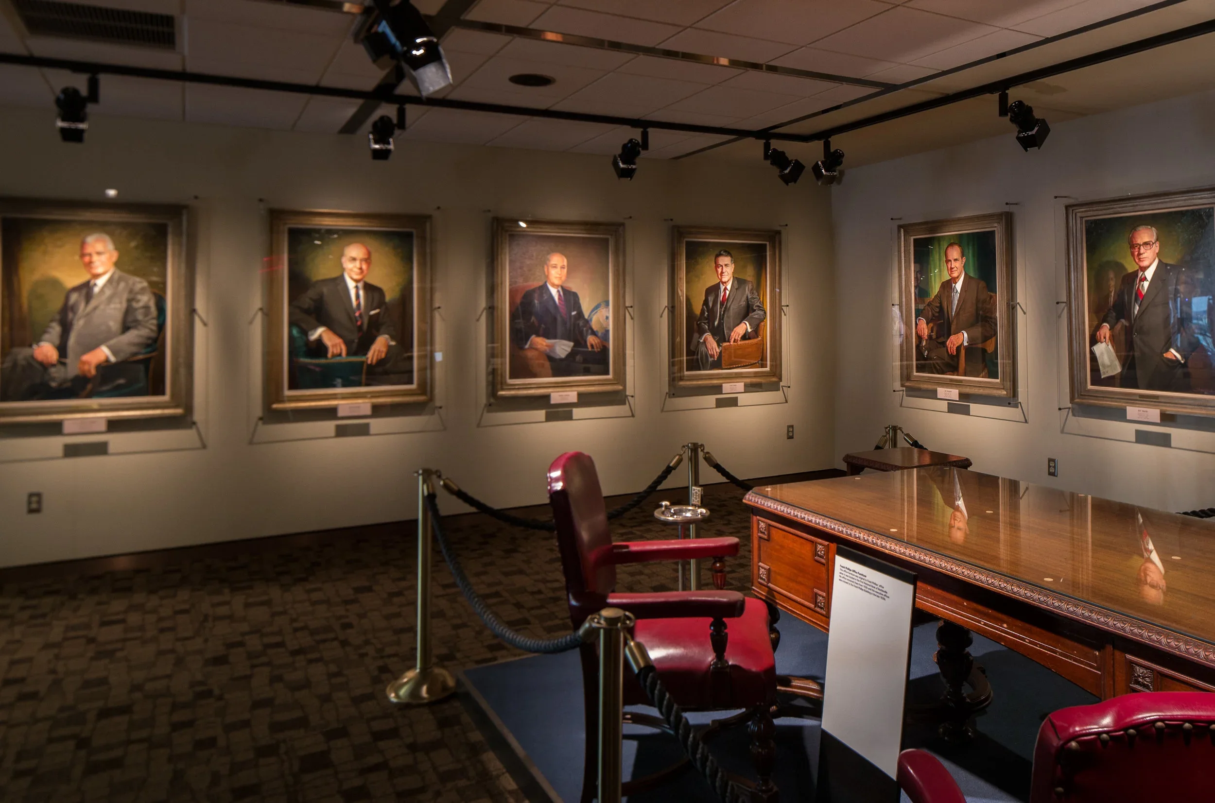 An art gallery exhibit showcasing six large portraits of distinguished men dressed in suits, hanging on a beige wall, with a polished wooden desk and burgundy chairs in the foreground.