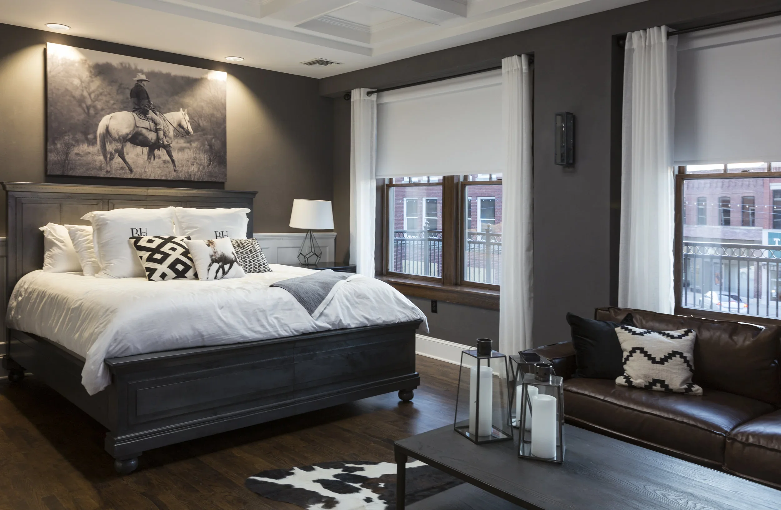 A bedroom with a large bed, black wooden headboard, white bedding, and decorative pillows. There is a black and white cowhide rug and a brown leather sofa with a patterned pillow. Three candles in glass holders are on a coffee table. The room has two