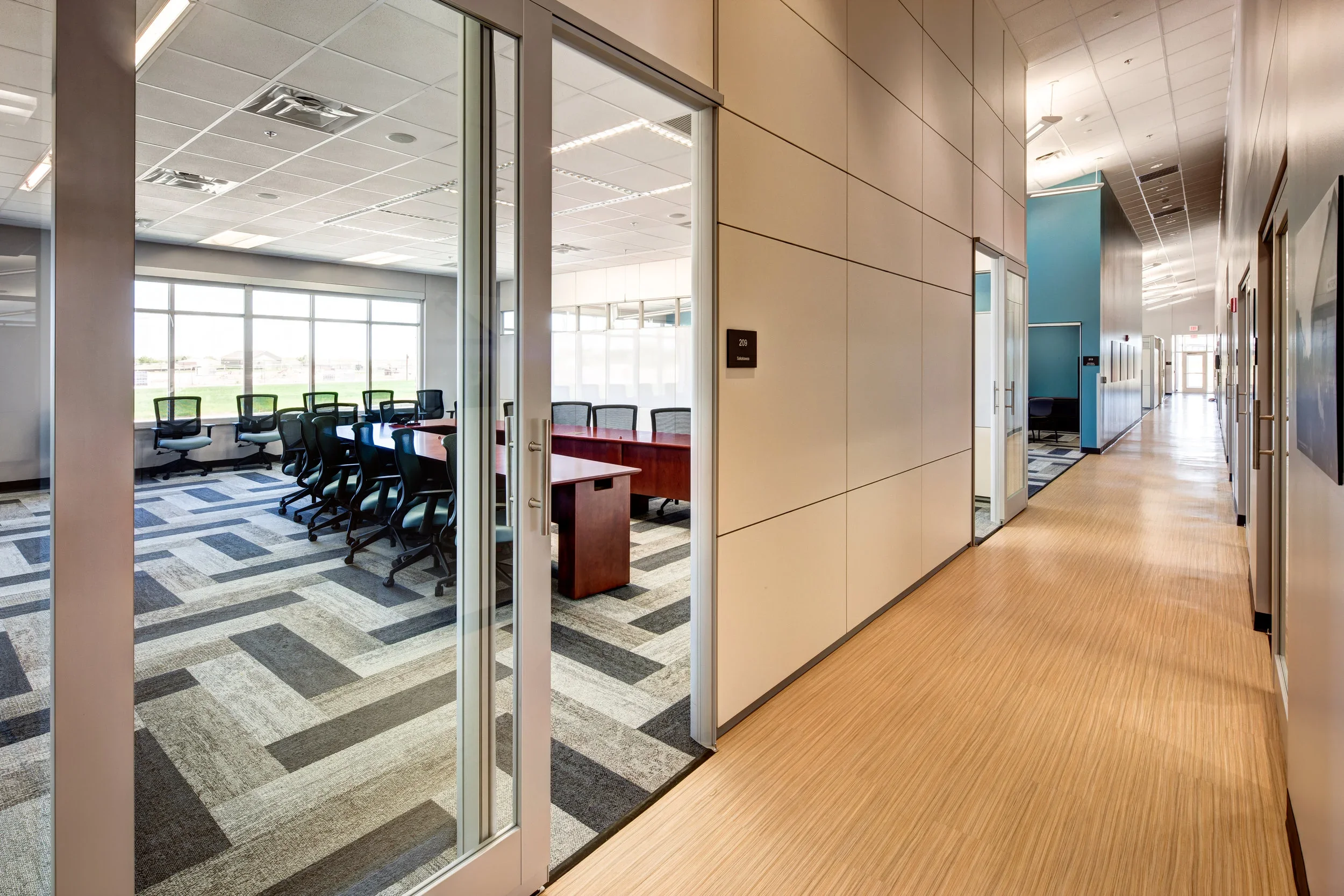Empty office corridor with beige walls and wooden flooring, leading to a conference room with large windows and chairs around a table.