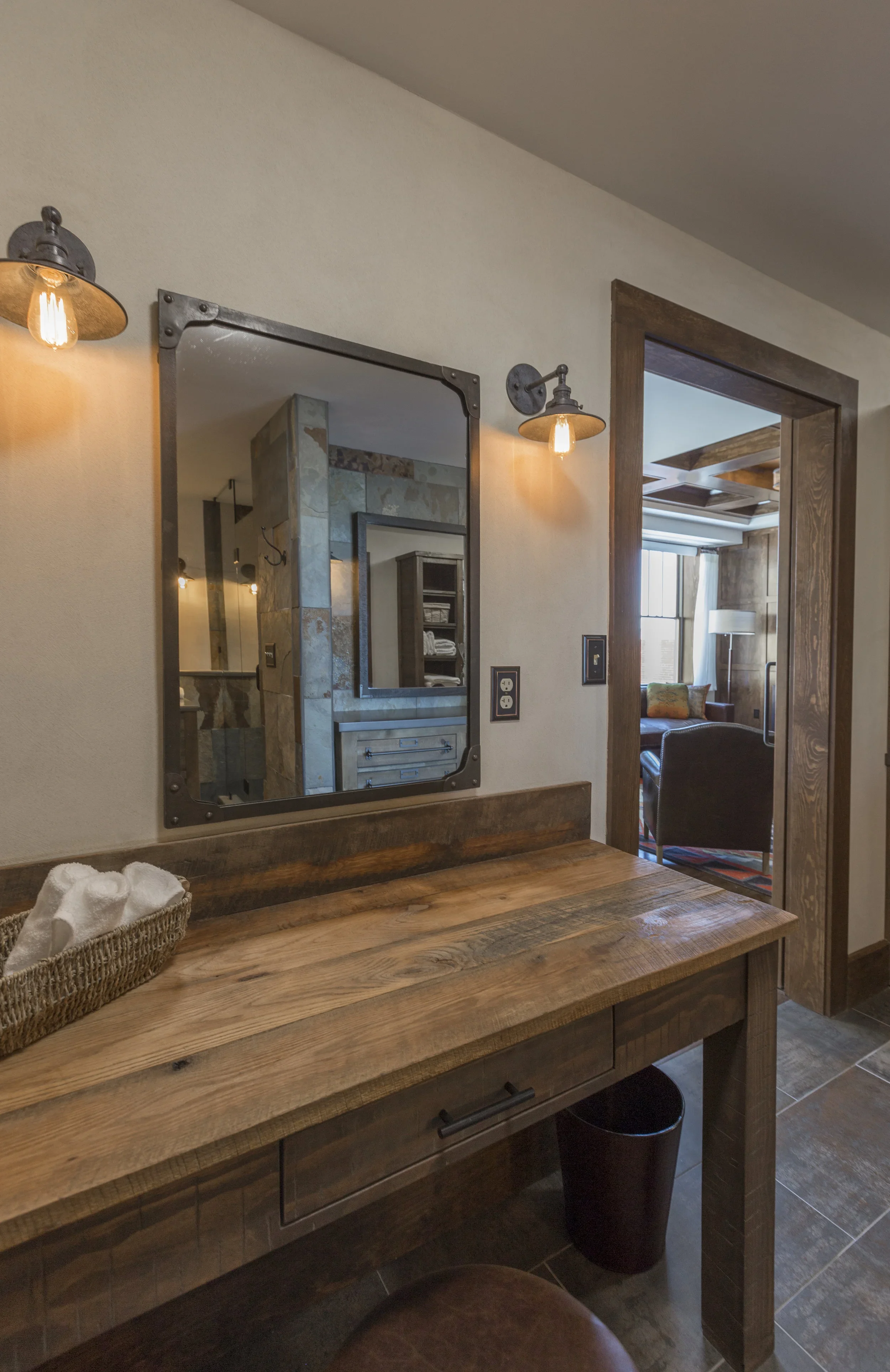 Interior of a rustic-style room with a wooden vanity, mirror, and wall-mounted light fixtures; visible through the doorway are a sitting area with a chair, window, and decorative wood paneling.