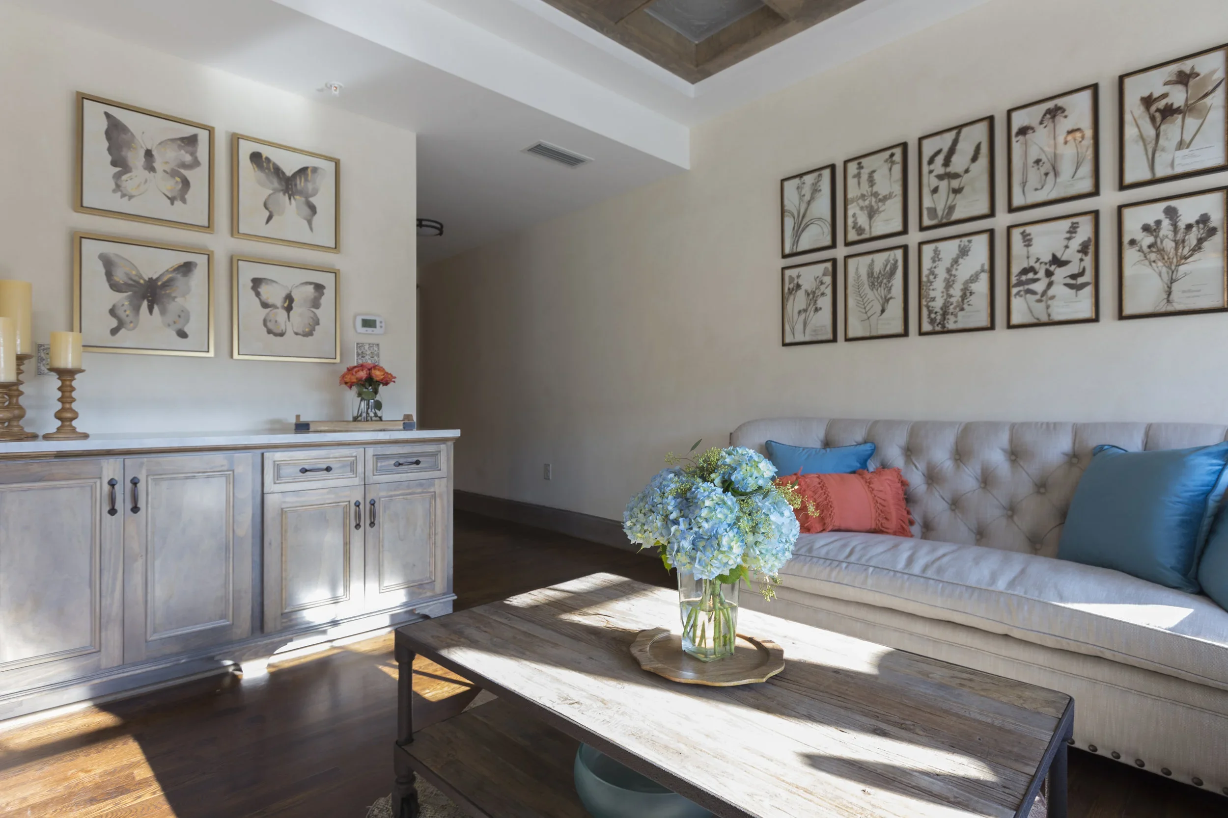Bright living room with a beige tufted sofa, colorful throw pillows, a wooden coffee table with a blue hydrangea flower arrangement, wall art of butterflies and pressed flowers, and a white sideboard with candles and a smaller flower arrangement.