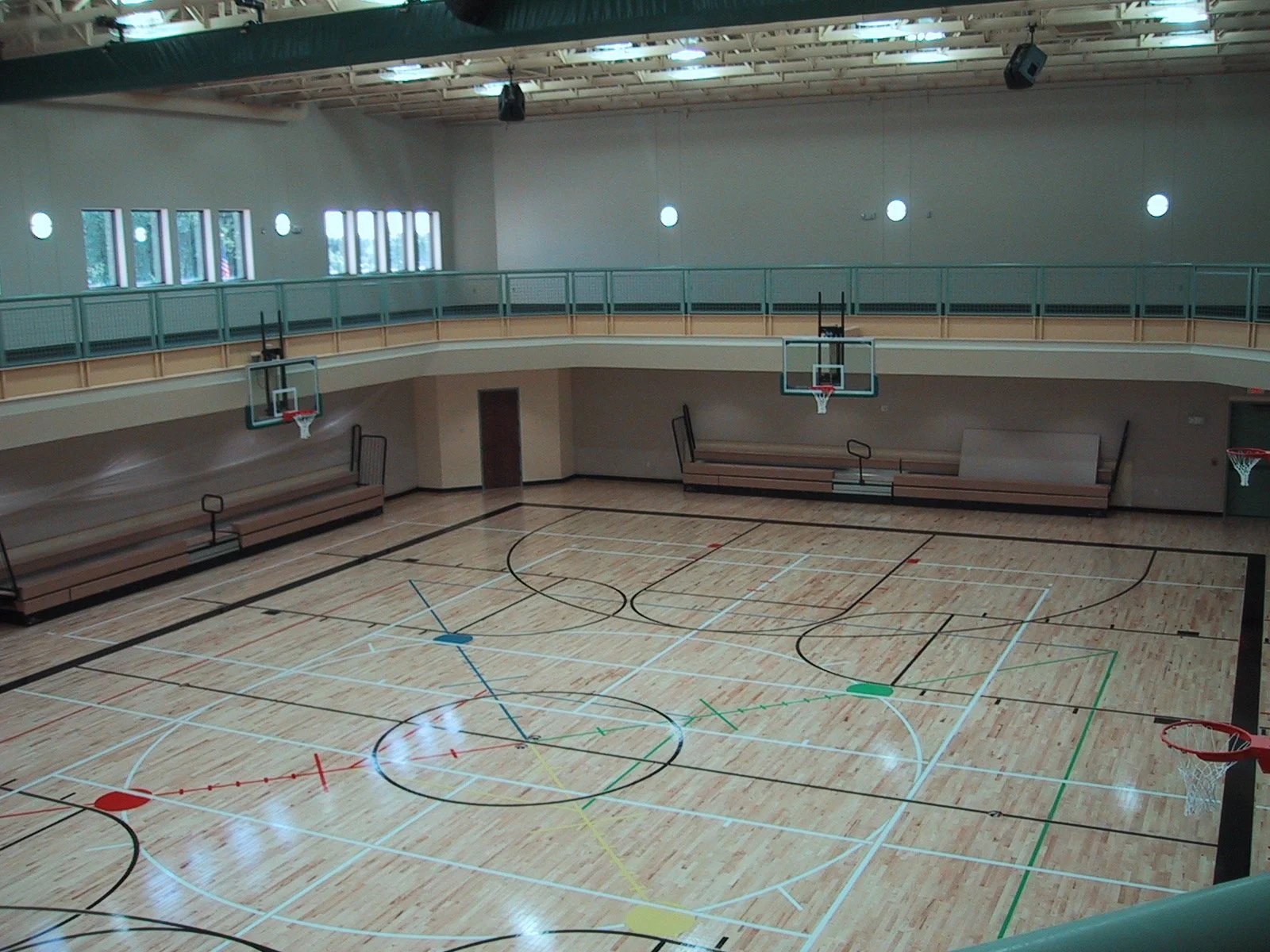 Indoor basketball court with wooden flooring, six basketball hoops, and benches along the walls, with windows and a balcony overlooking the court.