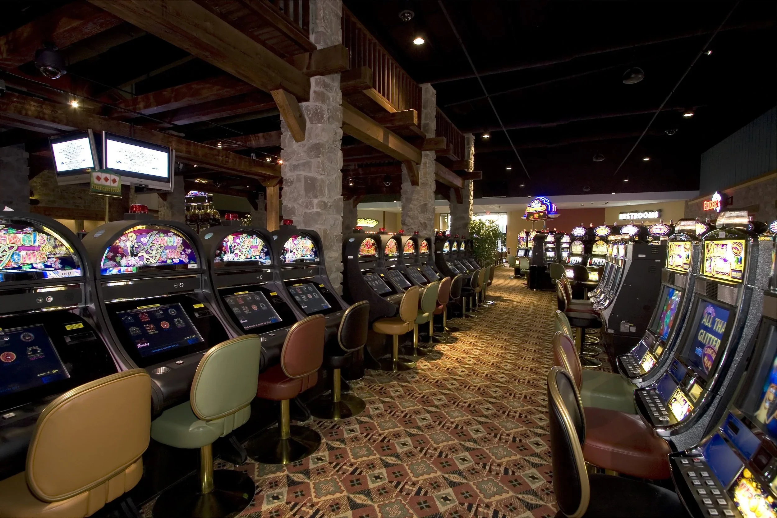 Inside a casino with rows of slot machines and chairs, stone pillars, wooden beams, and digital screens hanging from the ceiling.