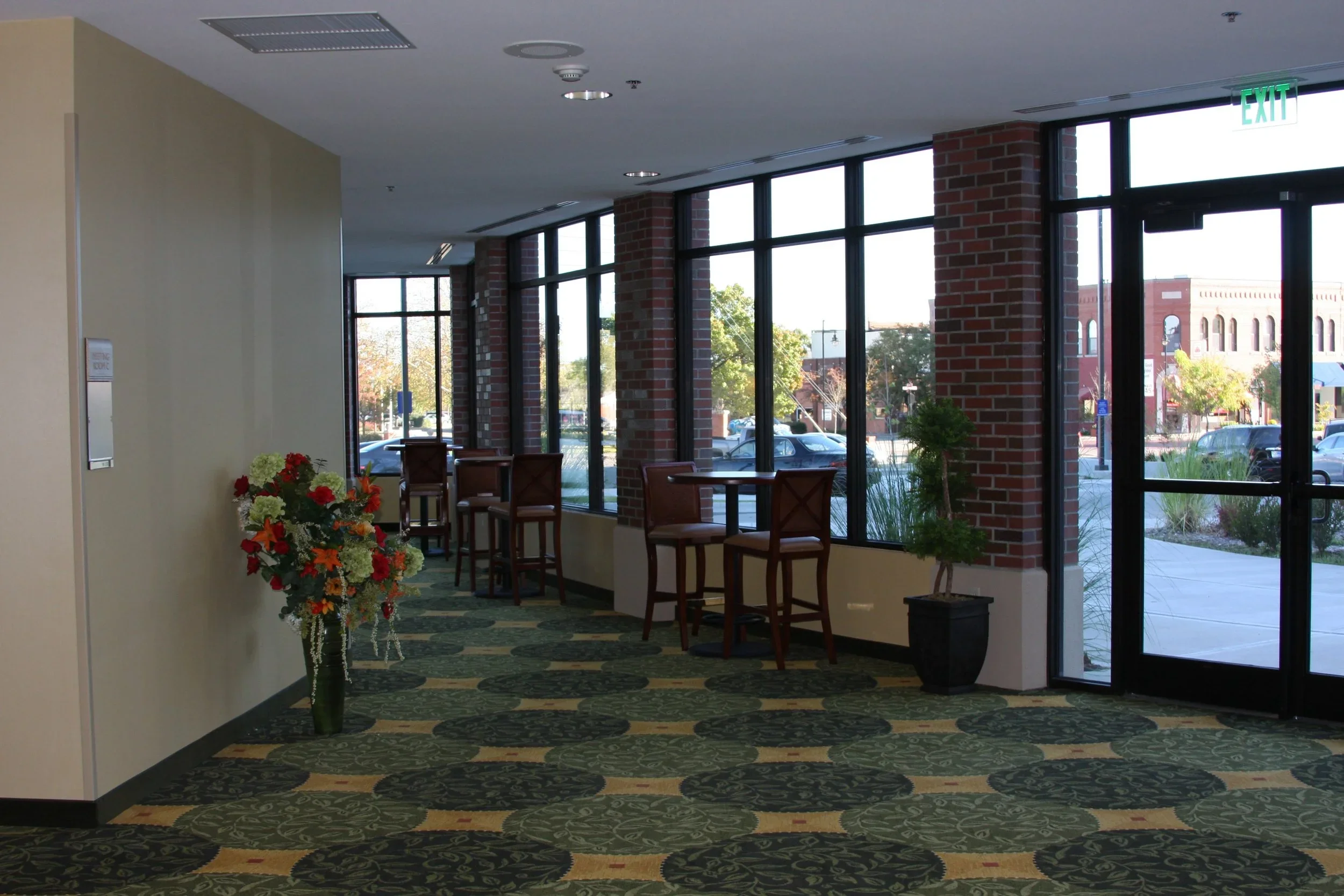 An empty indoor seating area with tall windows, potted plants, a floral arrangement, and a carpet with green and beige patterns.