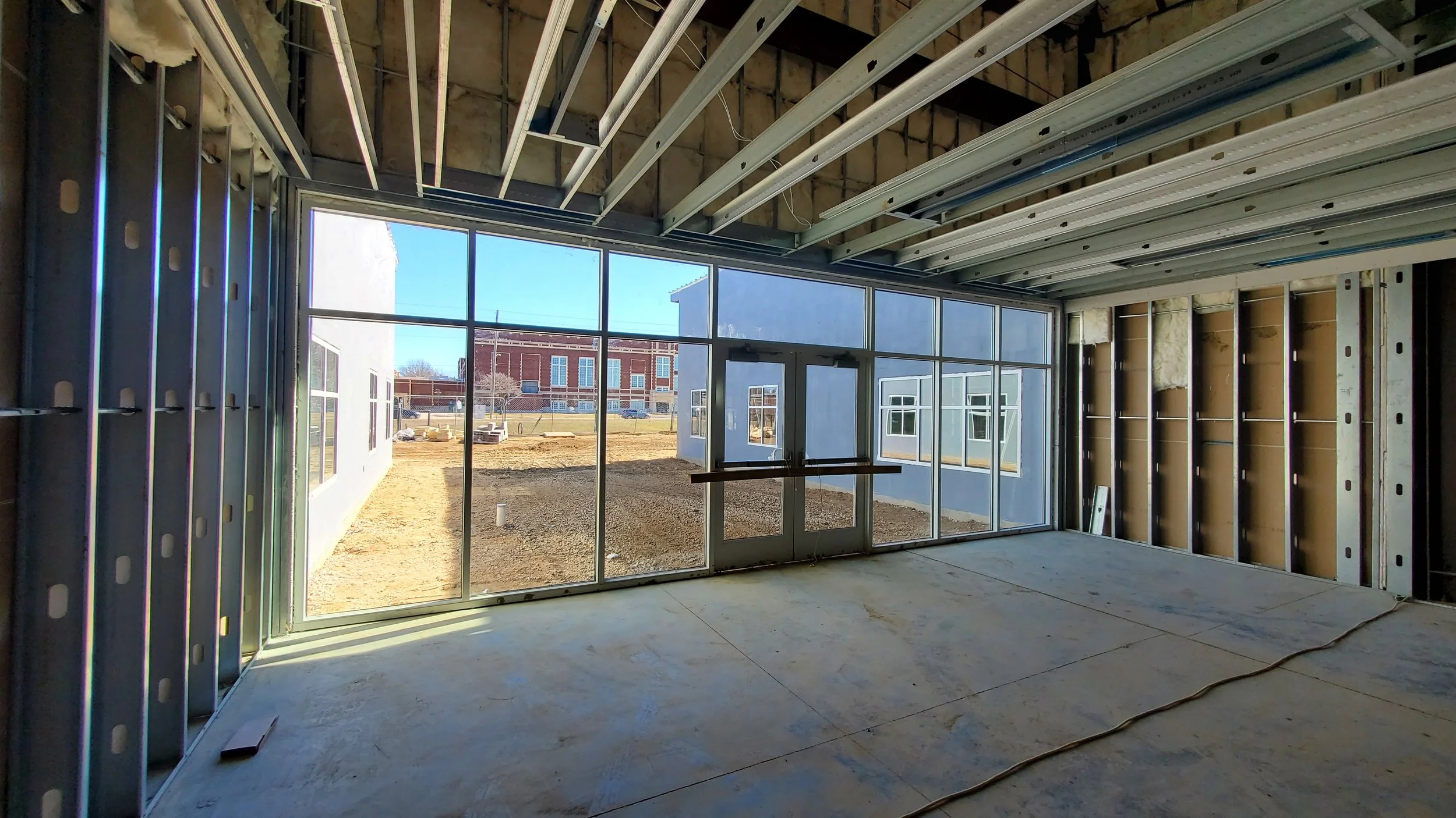 Interior view of a building under construction with metal framing, large glass windows, and unfinished walls, overlooking an outdoor area with dirt ground and nearby buildings.