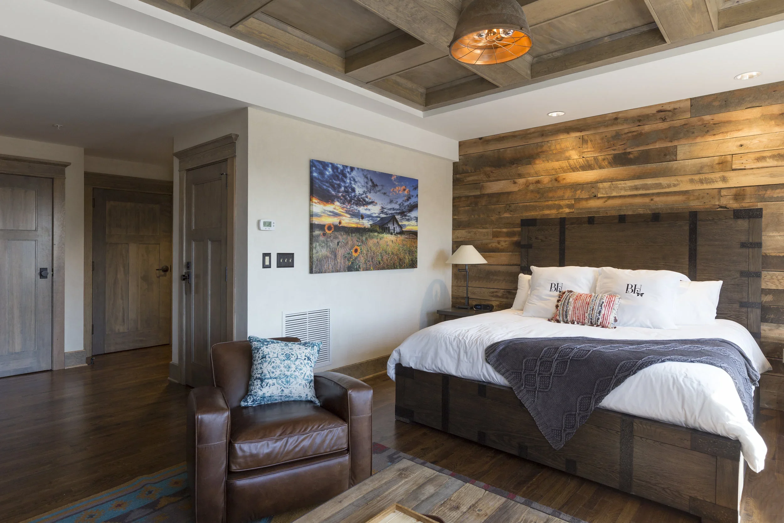 Modern bedroom with wooden accent wall, large bed with white bedding and decorative pillows, leather armchair with a blue pillow, and a landscape painting on the wall.