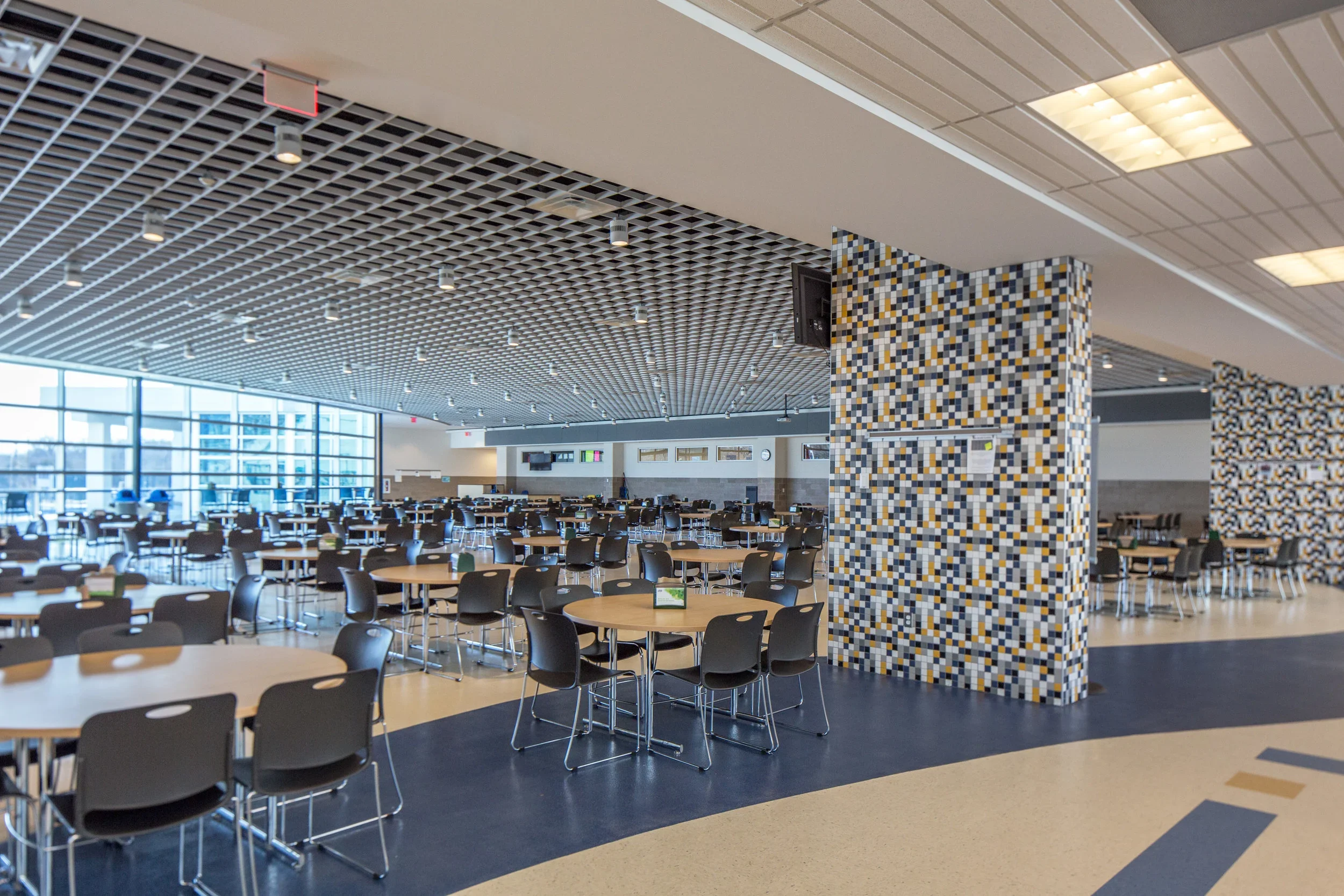A spacious cafeteria with round tables and black chairs, large windows letting in natural light, a decorative mosaic wall, and a grid ceiling with multiple light fixtures.