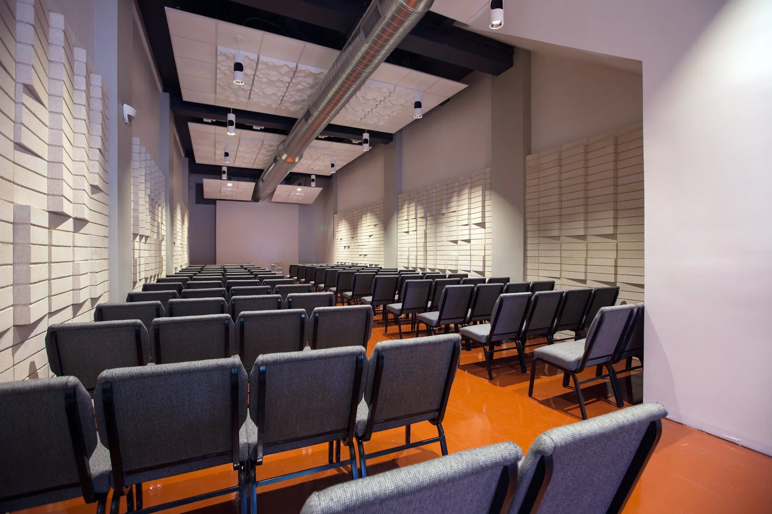View of an empty conference room with rows of black chairs facing a wall and a large screen, high ceiling with exposed ductwork, and acoustic panels on the walls.
