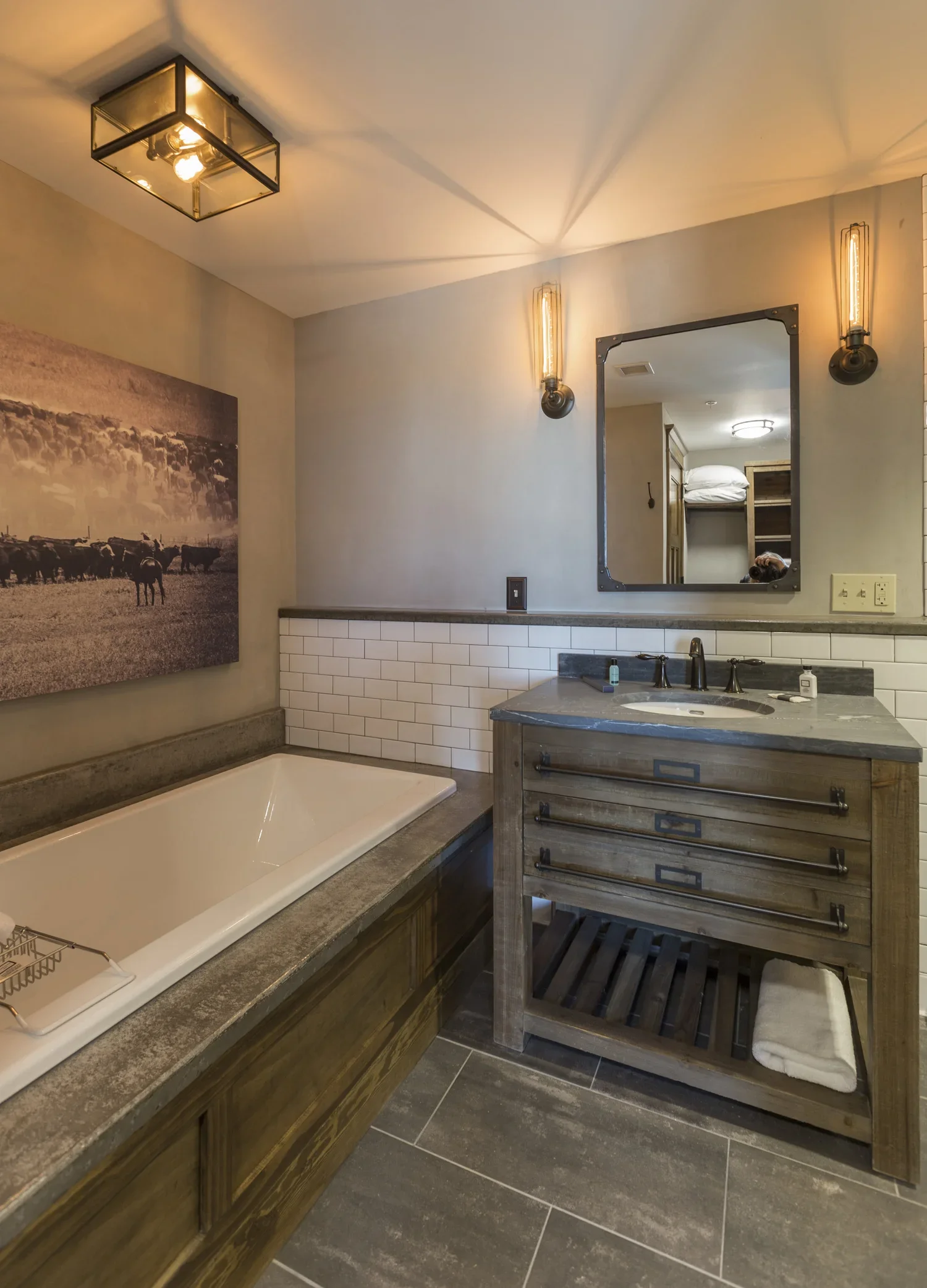 Modern bathroom with a bathtub, wooden vanity, and mirror. Decor includes wall lights, artwork of cattle, and gray tile flooring.