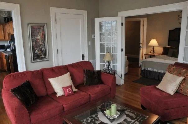 Living room with red sofas, pillows, a wooden coffee table, and a view into a bedroom with a nightstand and lamp.