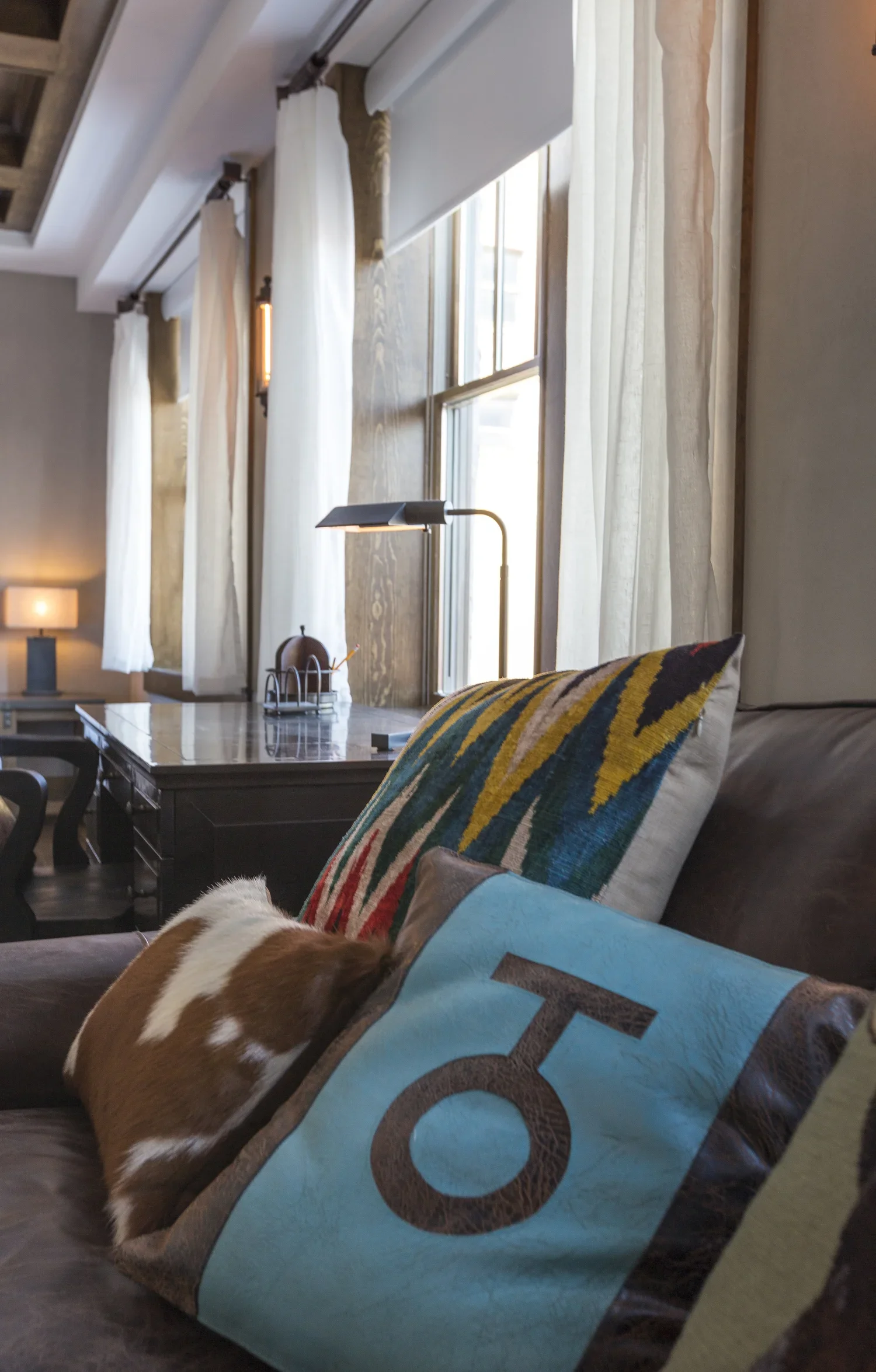 Living room with white curtains, a desk near the window, and decorative pillows on a sofa; one pillow with a colorful pattern and another with a blue and brown design featuring a horseshoe symbol.