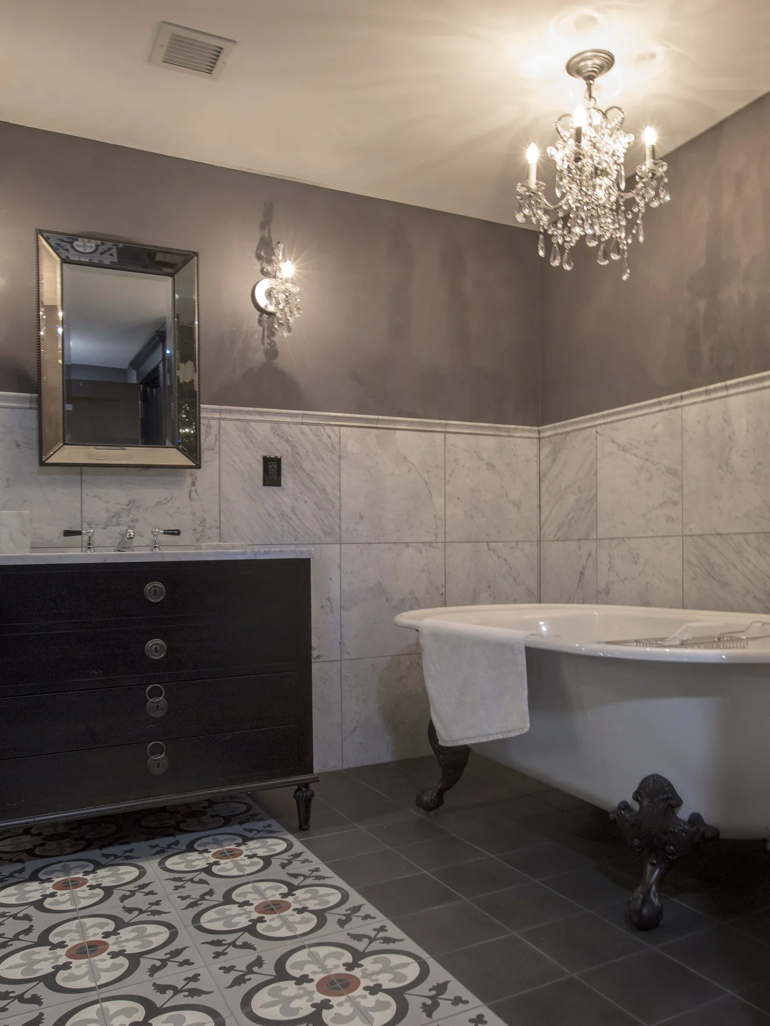 Elegant bathroom with dark vanity, black clawfoot bathtub, patterned tile floor, grey walls, crystal chandelier, and mirror.