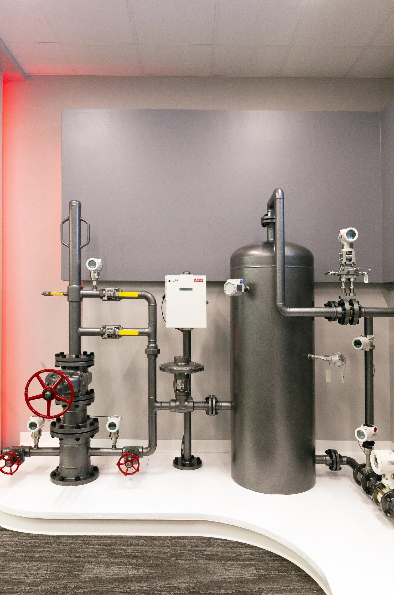 Industrial piping system with valves, gauges, and a large metal tank in a laboratory or facility setting.