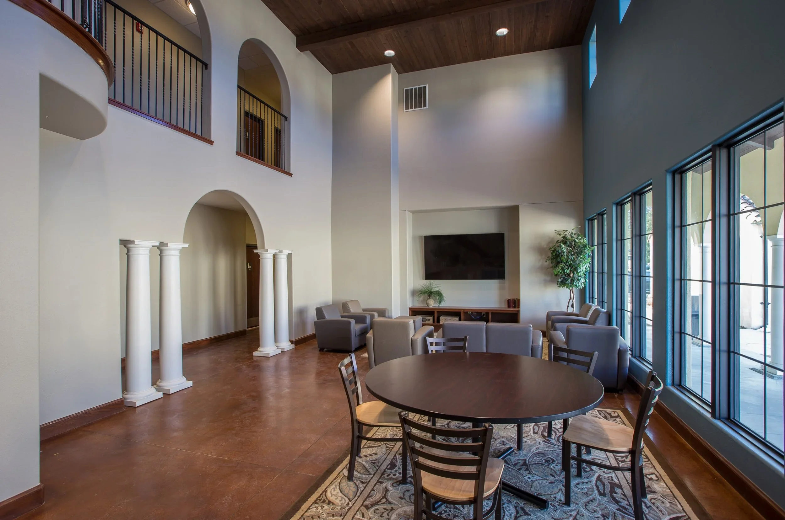 Spacious living room with high ceiling, large windows, and patio doors, furnished with a round dining table, six chairs, multiple armchairs, a large wall-mounted television, and potted plants.