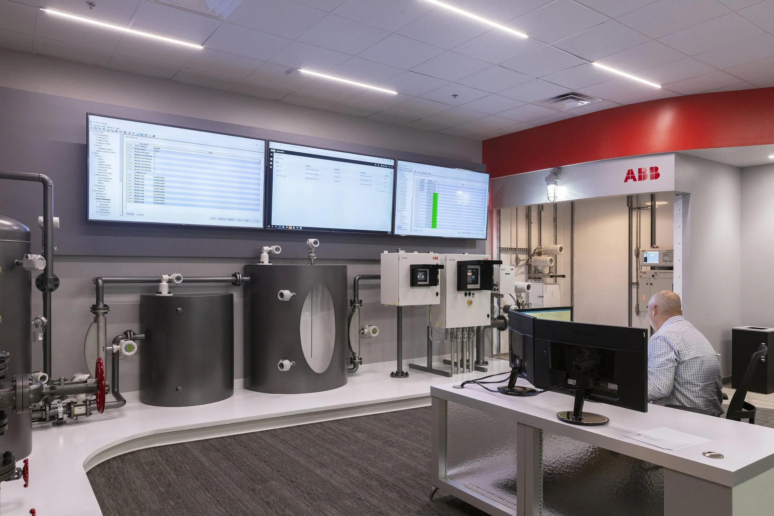 A control room with three large computer monitors displaying data, several industrial tanks and equipment, and a person working at a desk with dual monitors, all within an ABB facility.