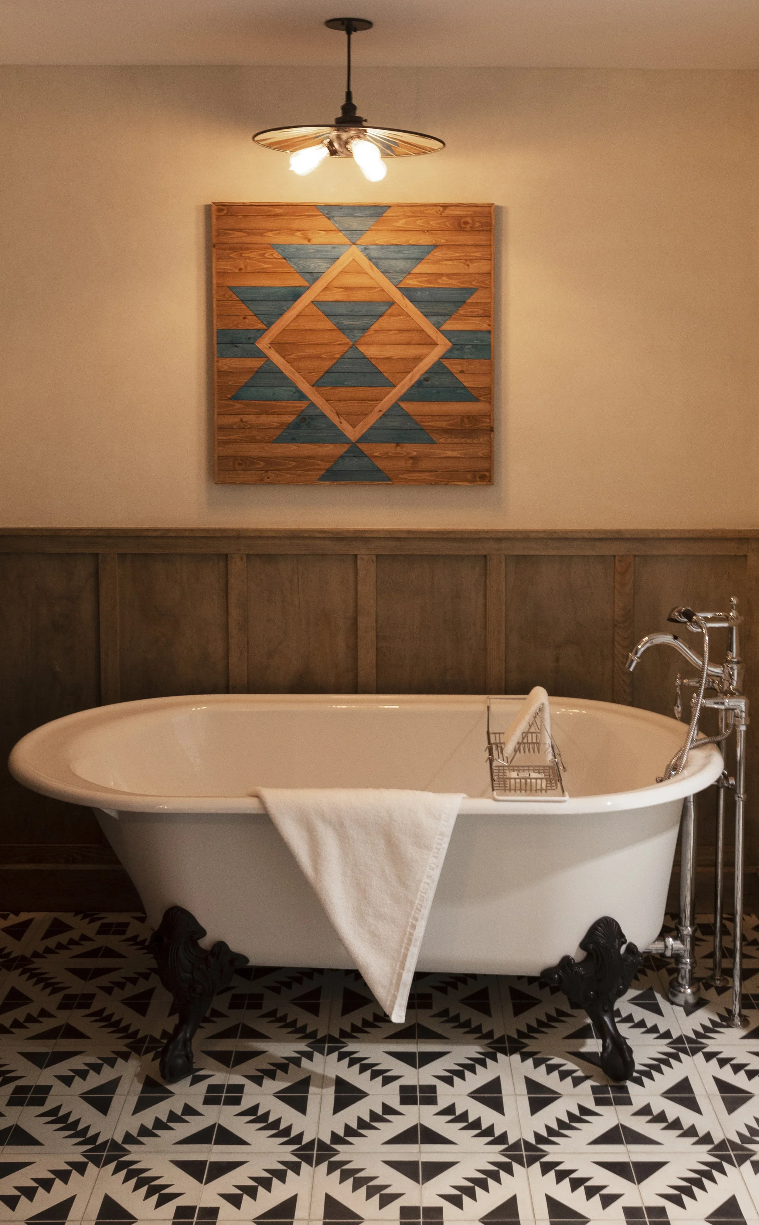 A vintage clawfoot bathtub with a white towel hanging over the side, set against a wall with wooden wainscoting, topped with a geometric wooden wall art piece, and illuminated by a modern ceiling light fixture, with black and white patterned tile flo