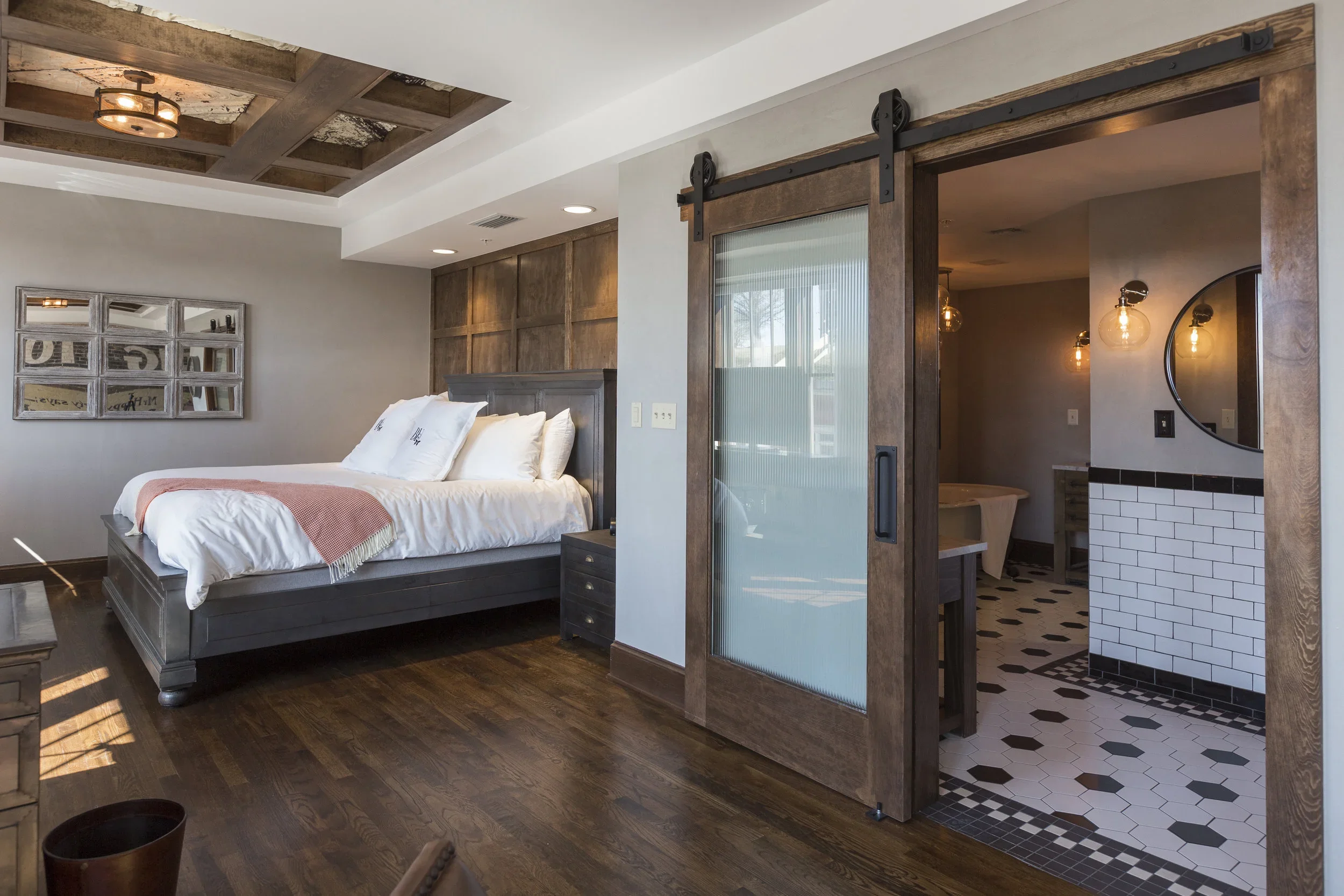 Bedroom with wooden floor, gray bed frame, white bedding, pink throw, and a sliding barn door leading to a bathroom with black and white hexagon tile floor, round mirror, and pendant lights.