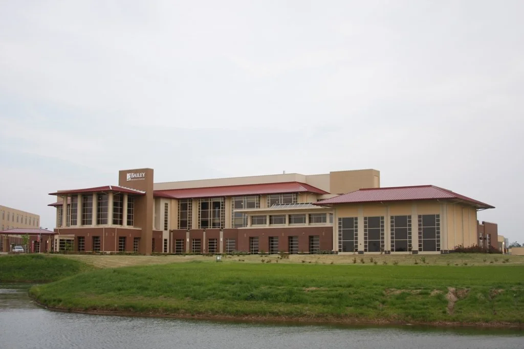Modern office building with a red roof, surrounded by green grass and a small body of water in the foreground.