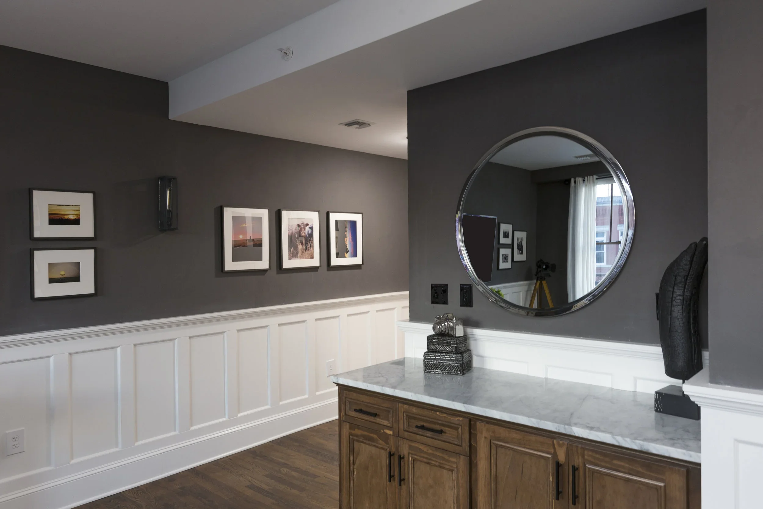 Interior of a modern room with a dark gray wall, a white half-wall wainscoting, a wooden cabinet with a marble countertop, a round mirror, framed photographs on the wall, and a window with curtains.