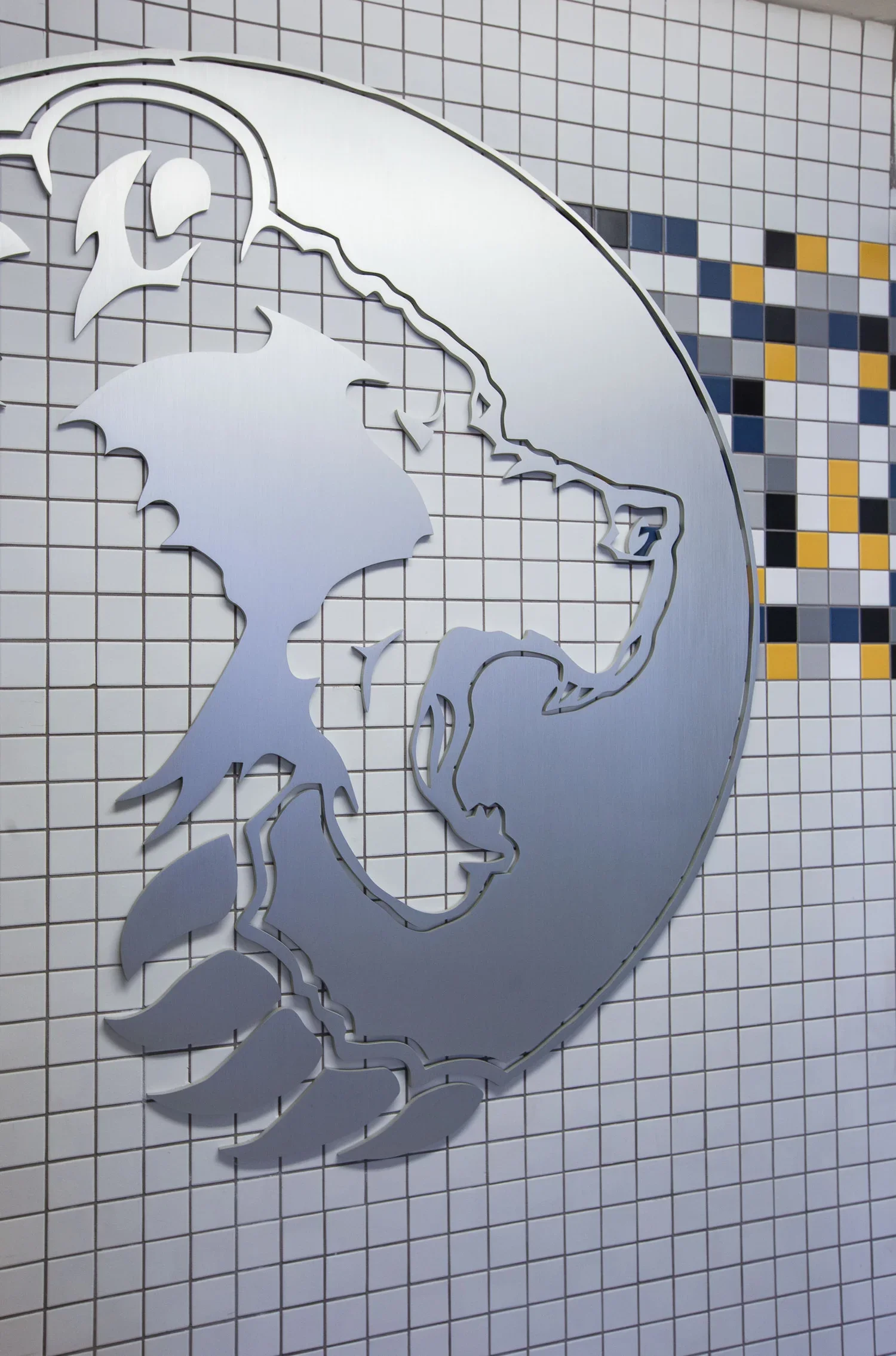 The image shows a metallic cutout of a globe mounted on a tiled wall. The globe depicts the continents of North and South America, with a background of small, multicolored tiles.