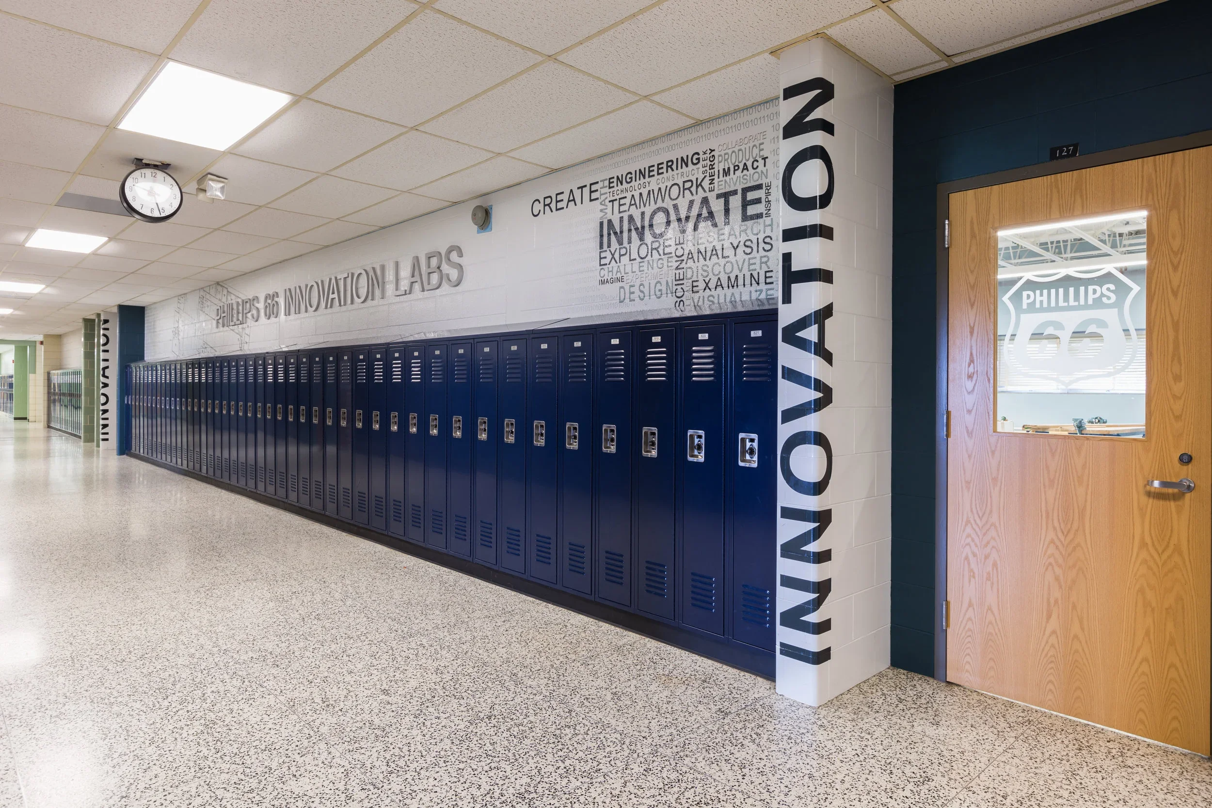 School hallway with blue lockers lining the wall, motivational words on the wall, a clock, and a door with a glass window leading to a room labeled Phillips 66.