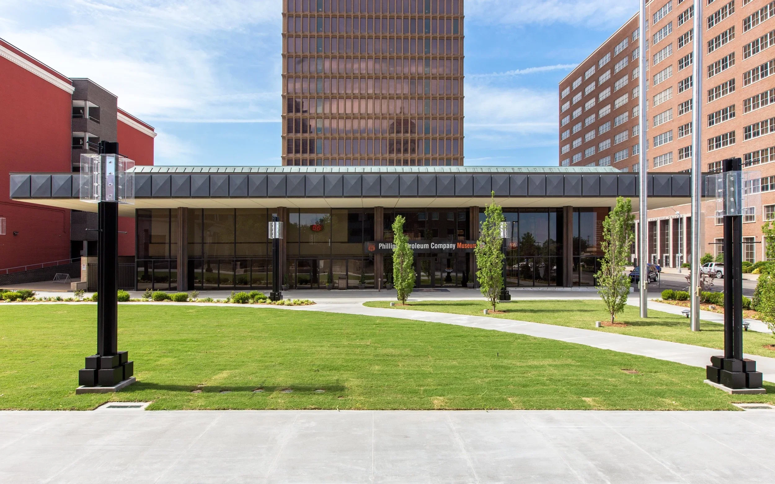 Exterior of the Phillips Petroleum Company Museum building with well-maintained lawn, young trees, and flagpoles in front, set against a backdrop of skyscrapers and a blue sky.