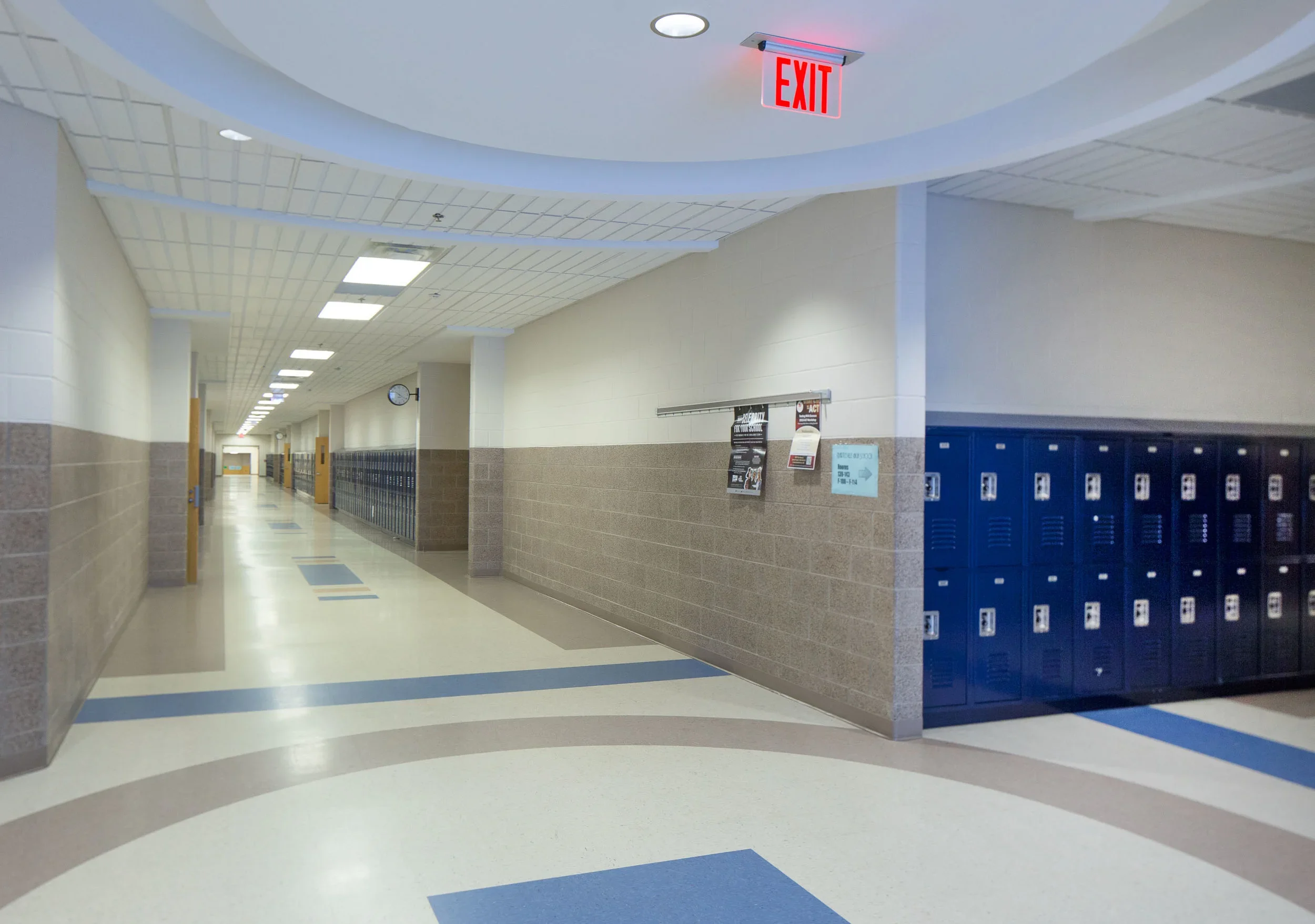 Empty school hallway with lockers on the right, clock on the wall, and an illuminated exit sign on the ceiling.
