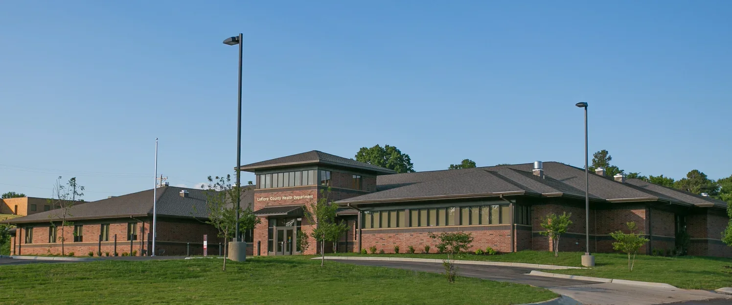Exterior view of LeFlore County Health Department building with brick walls, large windows, green grass, young trees, and street lamps under a clear blue sky.