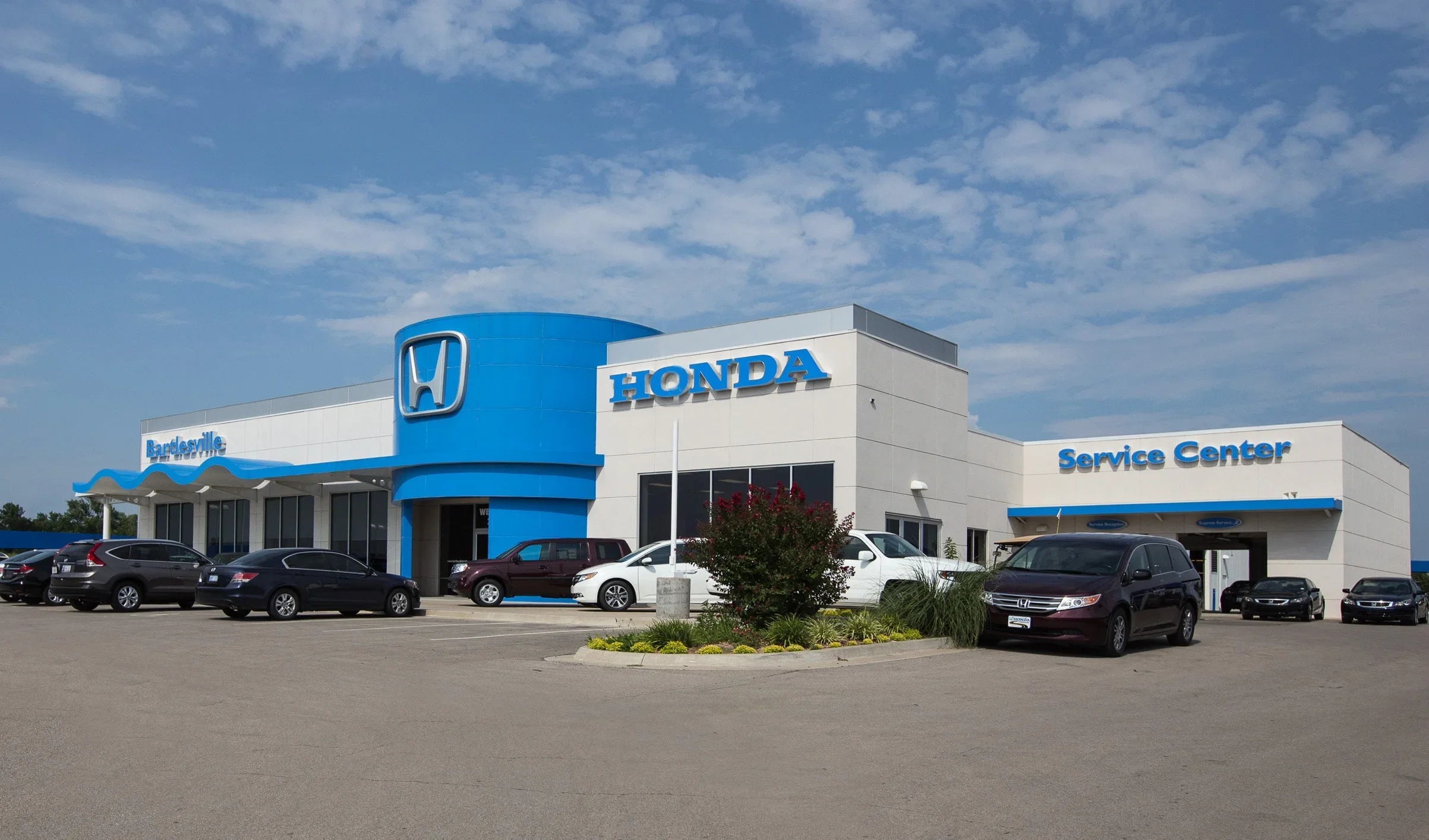 Exterior view of a Honda car dealership with a service center, several parked cars, a landscaped area with bushes and flowers, under a partially cloudy sky.