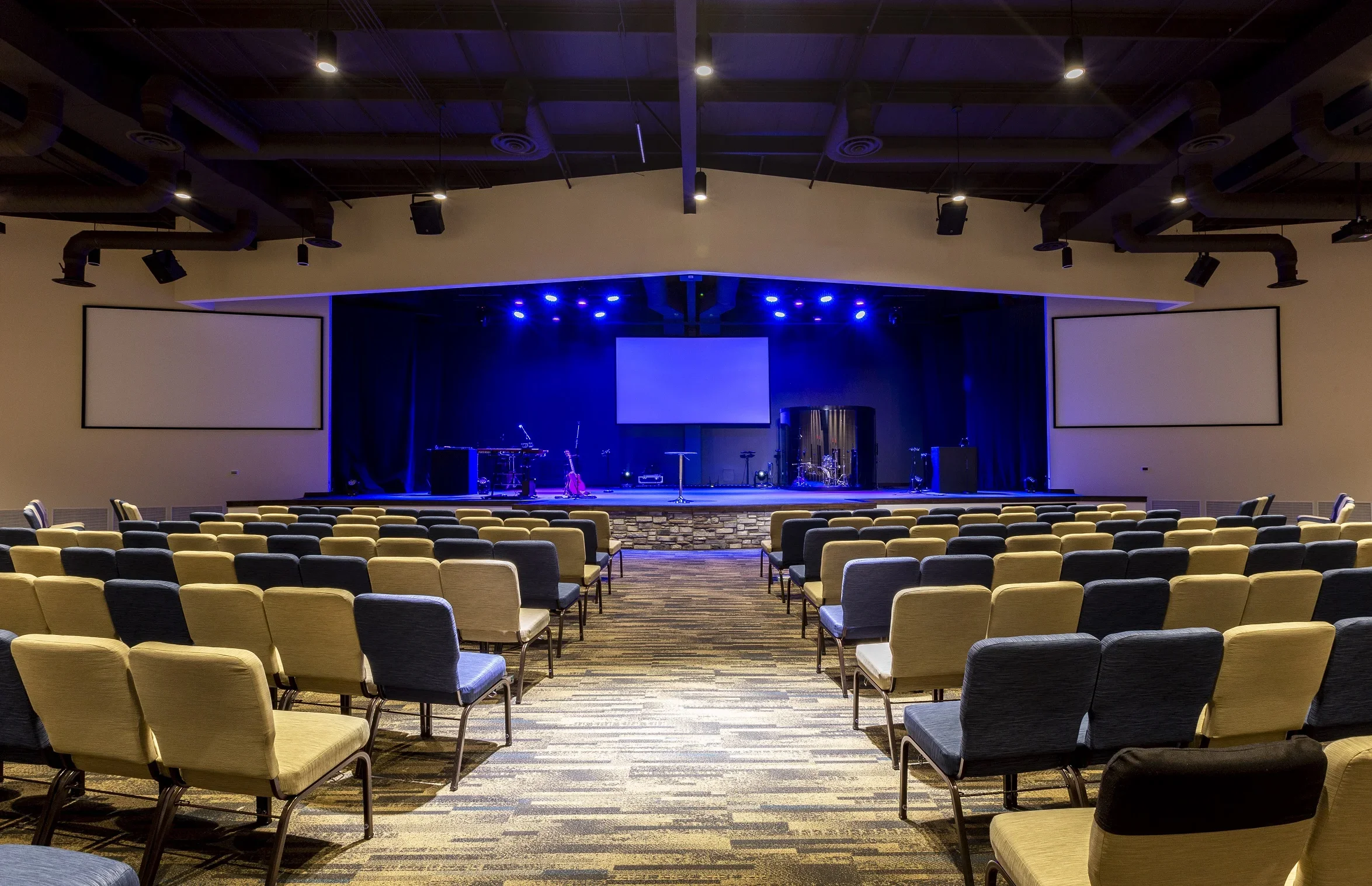 Empty auditorium with rows of chairs facing a stage with musical instruments and large screens, illuminated by blue stage lighting.