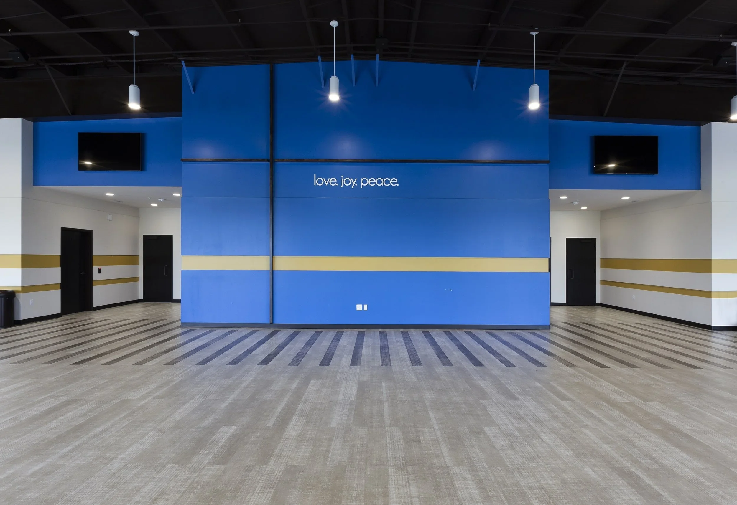A spacious, modern room with a blue accent wall that displays the words 'love. joy. peace.' in white text. The room has a beige and brown striped carpet, white walls with yellow horizontal stripes, two black doors on either side, and two large flat-s