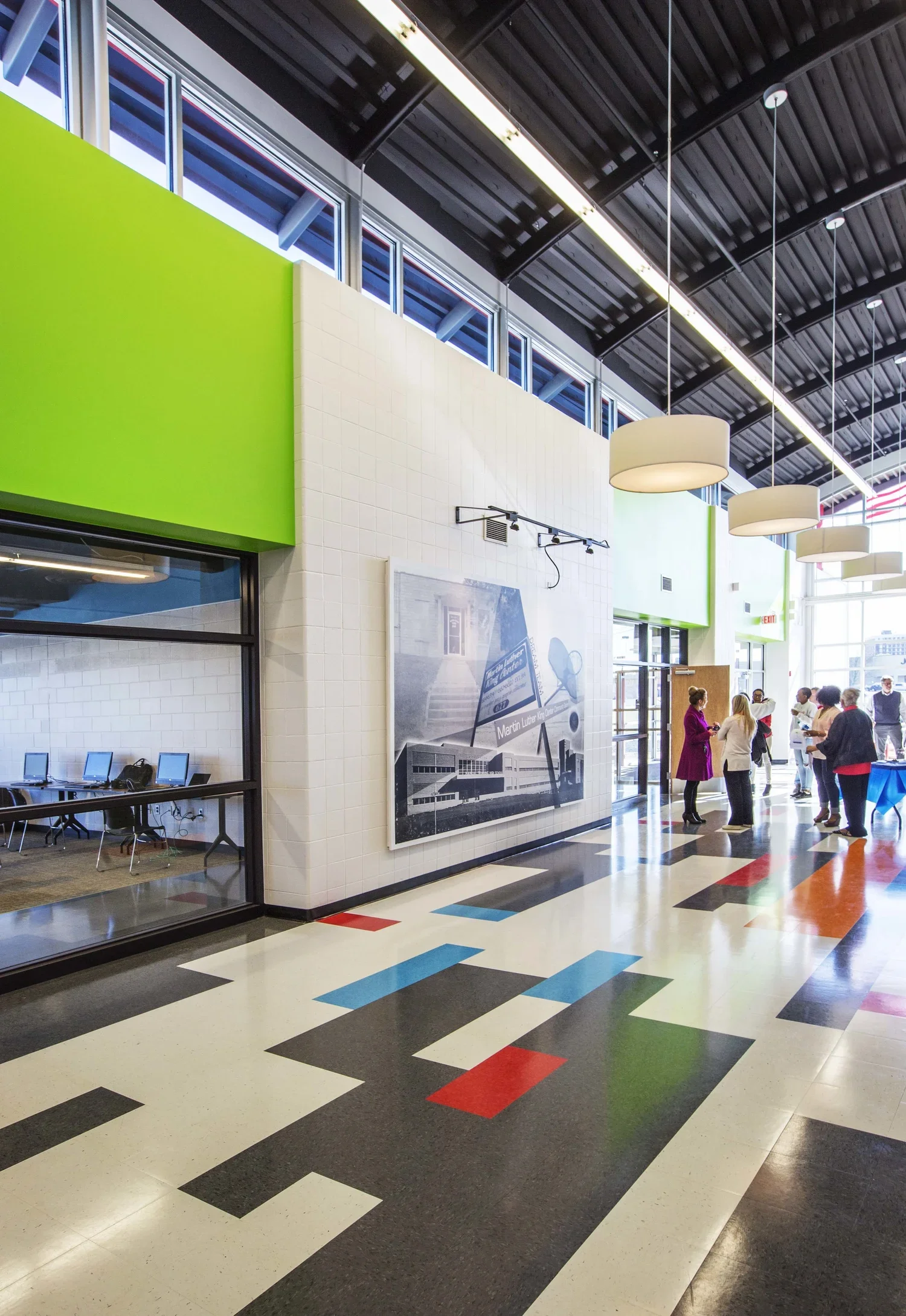 Indoor public space with colorful patterned floor, white tiled walls, green accents, large window, artwork on the wall, and a group of people standing and talking near the entrance.