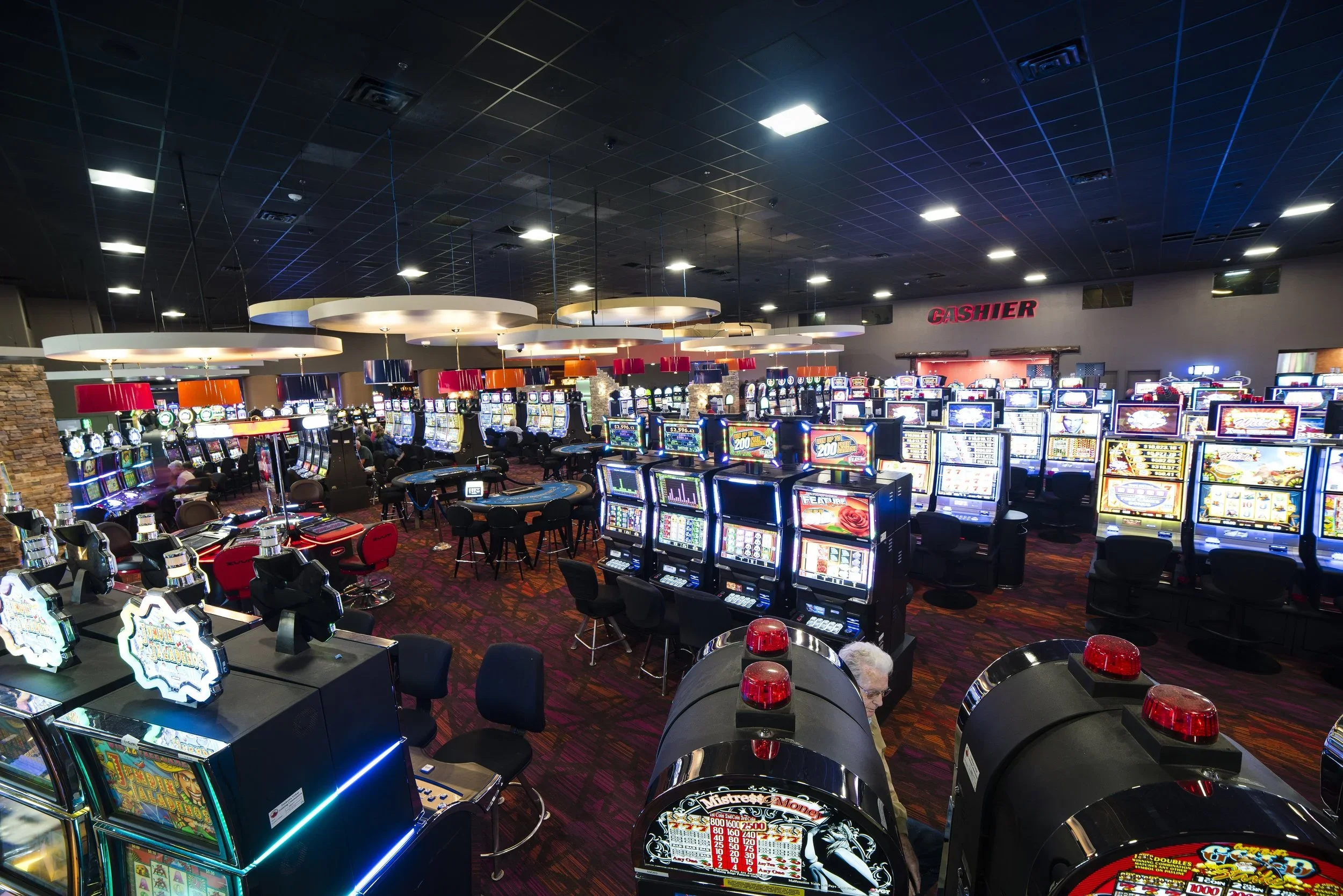 Inside a casino with rows of slot machines, a cash register sign, and people playing games.