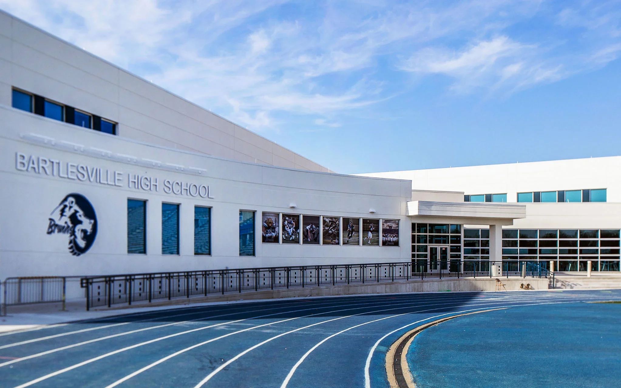 Exterior of Barlesville High School with a running track, windows, and school signs, under a blue sky with some clouds.