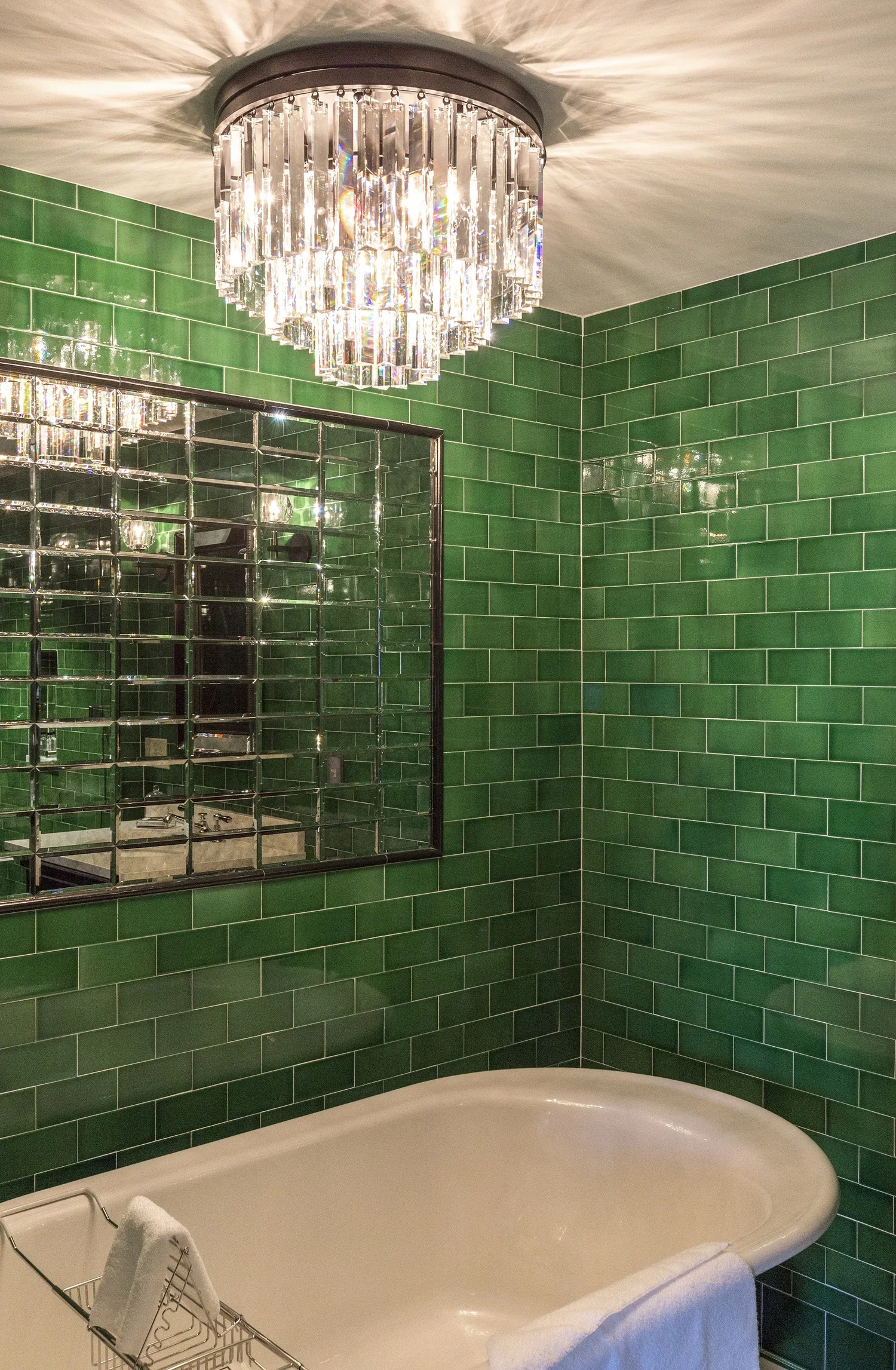 A bathroom with green tiled walls, a large mirror, a chandelier hanging from the ceiling, a white bathtub with a towel on its side, and a small metal rack holding a towel.