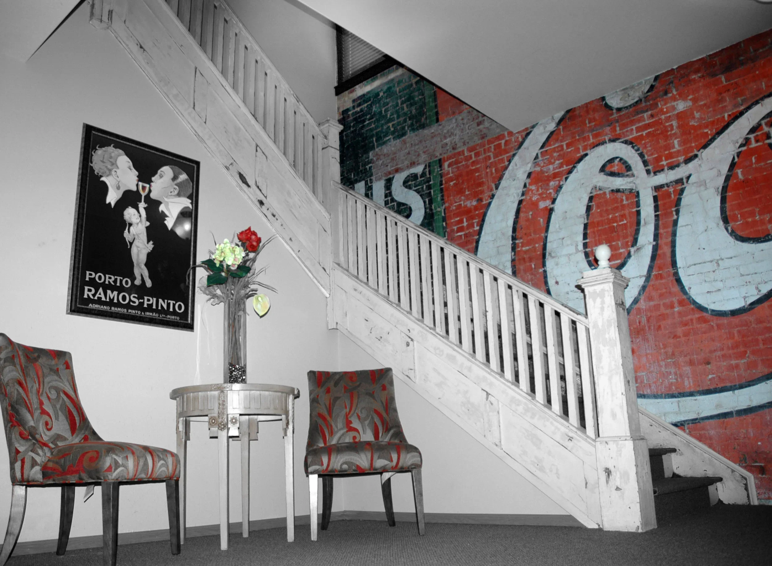 Interior of a vintage cafe or bookstore with a staircase, two patterned chairs, a small round table with a vase of flowers, a framed Porto Ramos-Pinto poster, and a large rustic Coca-Cola mural on exposed brick wall.